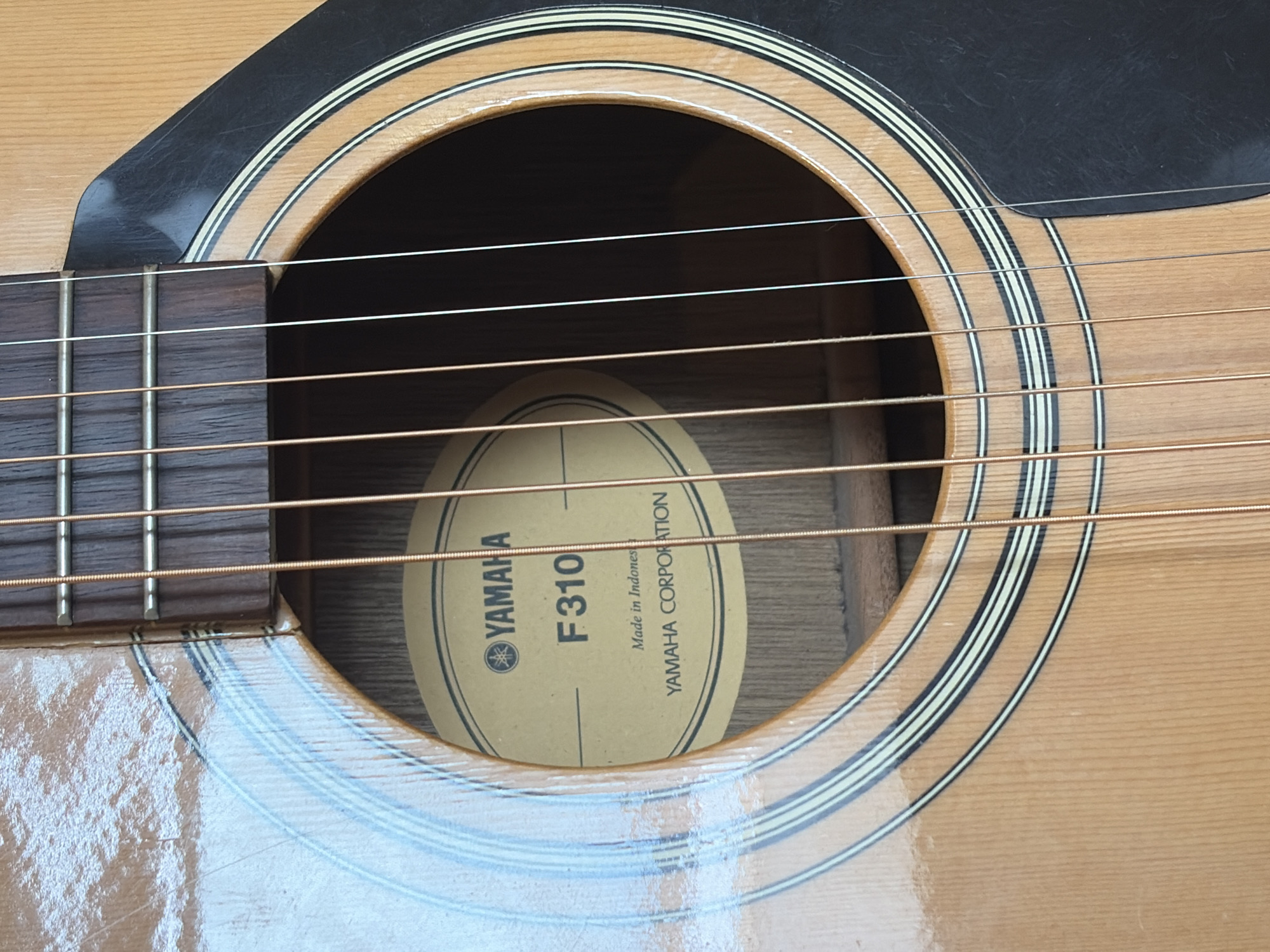 The image shows a close-up view of the soundhole and strings of an acoustic guitar. A round, yellow label with black text is visible inside the soundhole. The text on the label reads "YAMAHA" with a logo, "F310", "Made in Indones", and "YAMAHA CORPORATION". The guitar's body is made of wood with a glossy finish, and it has decorative rings around the soundhole. The strings appear to be made of metal. The scene is well-lit, with the lighting casting reflections on the guitar's surface. The background is dark, suggesting the guitar might be indoors or in a dimly lit environment. There are no people or animals in the image.