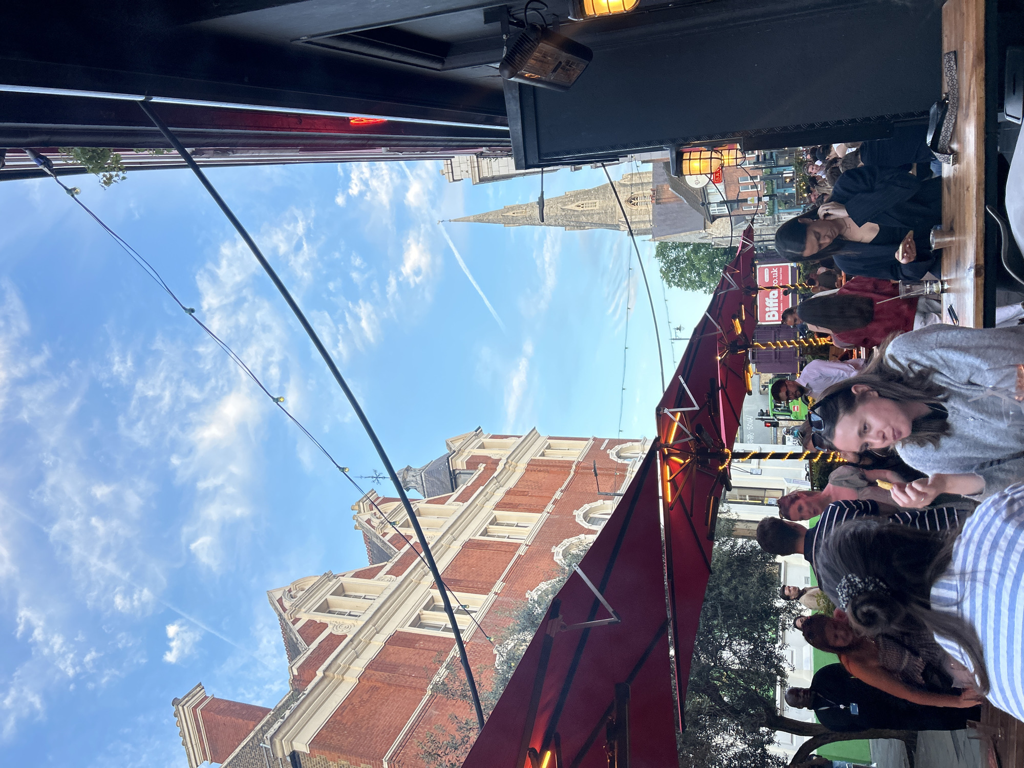 The image shows an outdoor dining scene in London, United Kingdom. A group of people are seated at tables under red awnings. They are engaged in conversation and appear to be enjoying a meal or drinks. In the background, a brick building and the spire of a church are visible against a blue sky with scattered clouds. Power lines run across the sky. The image is taken from an elevated position under a dark canopy, possibly inside a building.
