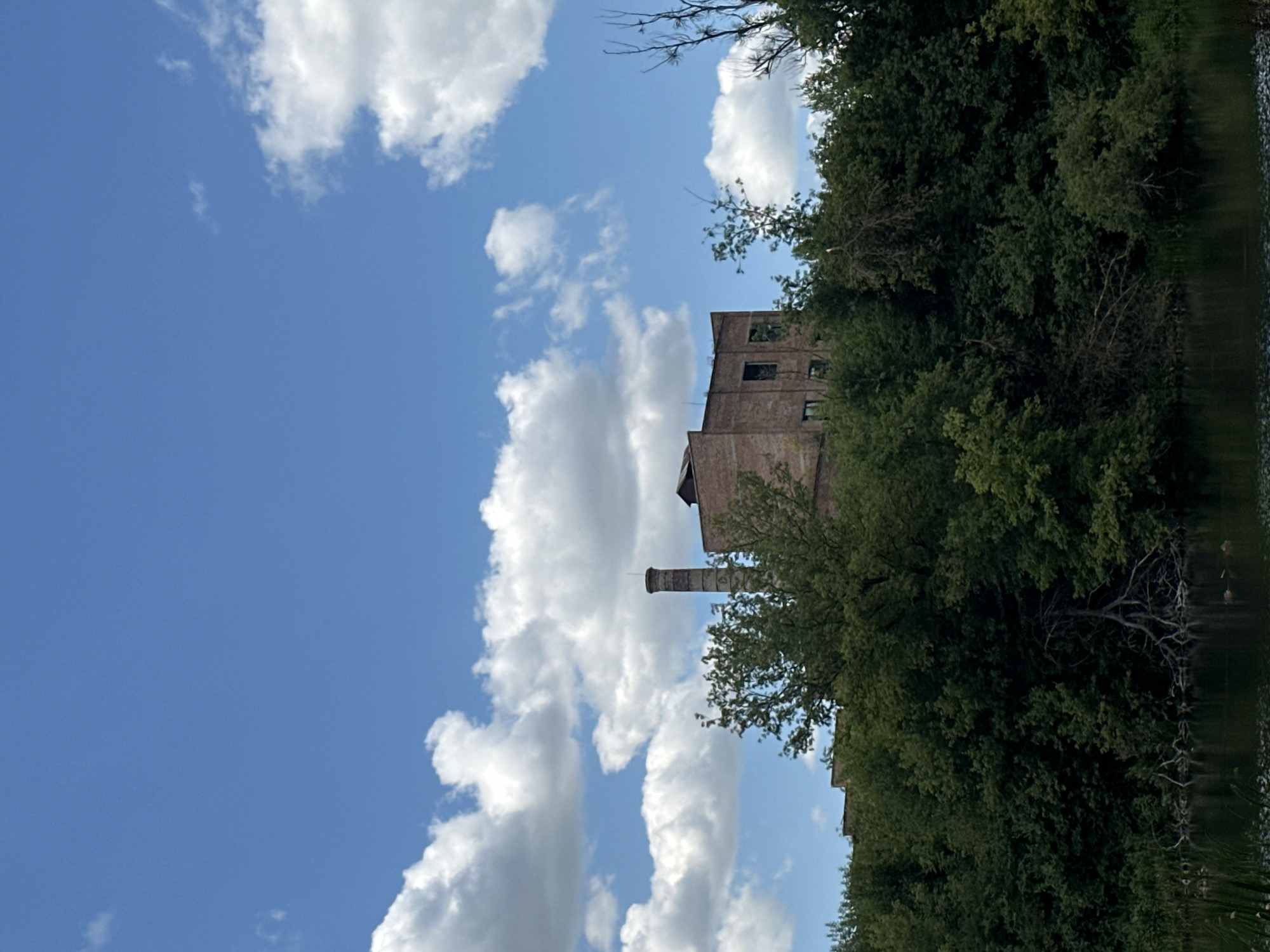 A long shot shows a brick factory and chimney peaking out from behind a dense line of trees. A calm body of water is in the foreground. The sky above is bright blue with scattered white clouds.