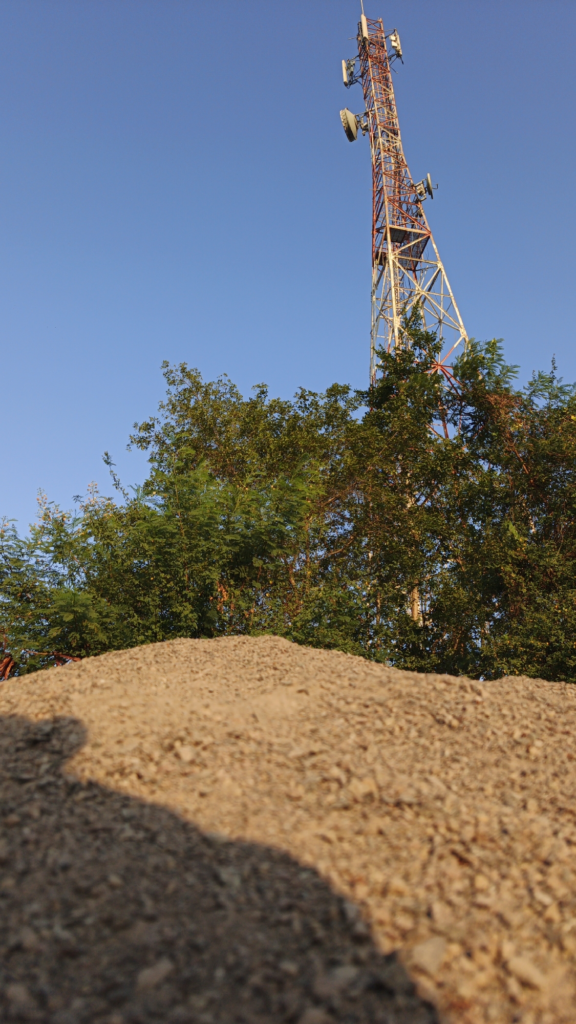 The image shows a tall telecommunications tower with multiple antennas, rising above a canopy of green trees against a clear blue sky. In the foreground, a mound of light brown gravel and soil is visible, with a shadow cast across it. The perspective is low-angle, emphasizing the height of the tower and the texture of the ground. This scene is located in Santing, Indonesia.