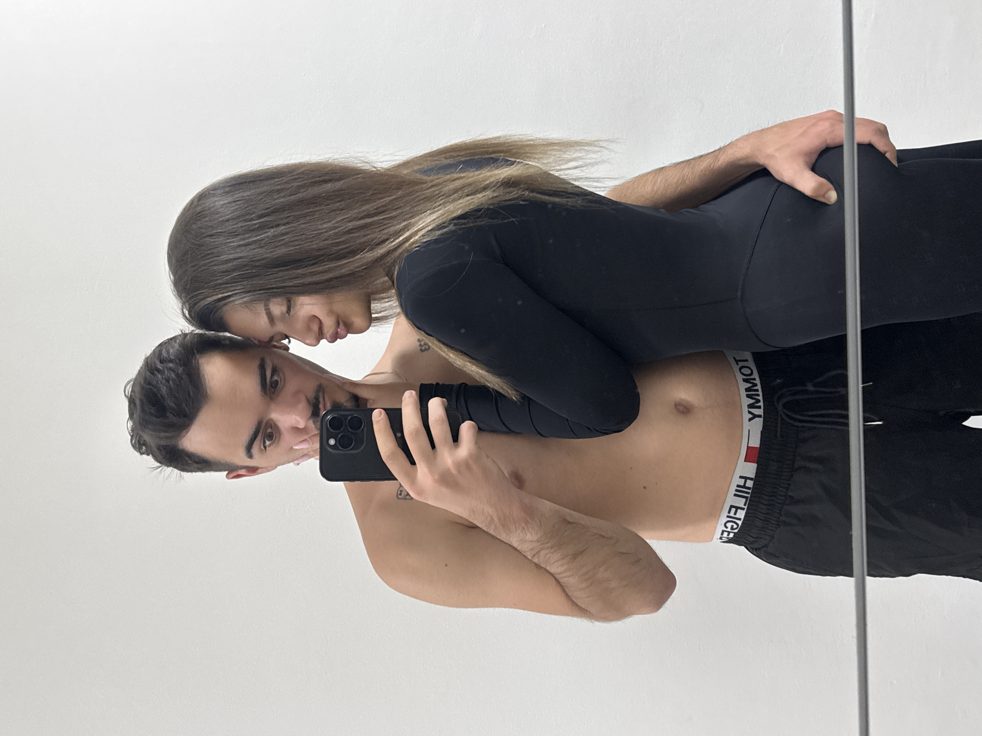 A waist-up indoor selfie taken in front of a mirror depicts a man and a woman. The man has dark hair, light skin, and a trimmed mustache, wearing black shorts with a Tommy Hilfiger waistband. He's shirtless, holding a black phone in his right hand to capture the image. The woman has long brown hair and is wearing a black bodysuit. She is positioned behind the man, with her left hand on his right shoulder and her right hand on his hip. There's a visible tattoo on her left collarbone. The background is a plain white wall. The location is not identifiable as Sofia, Bulgaria.