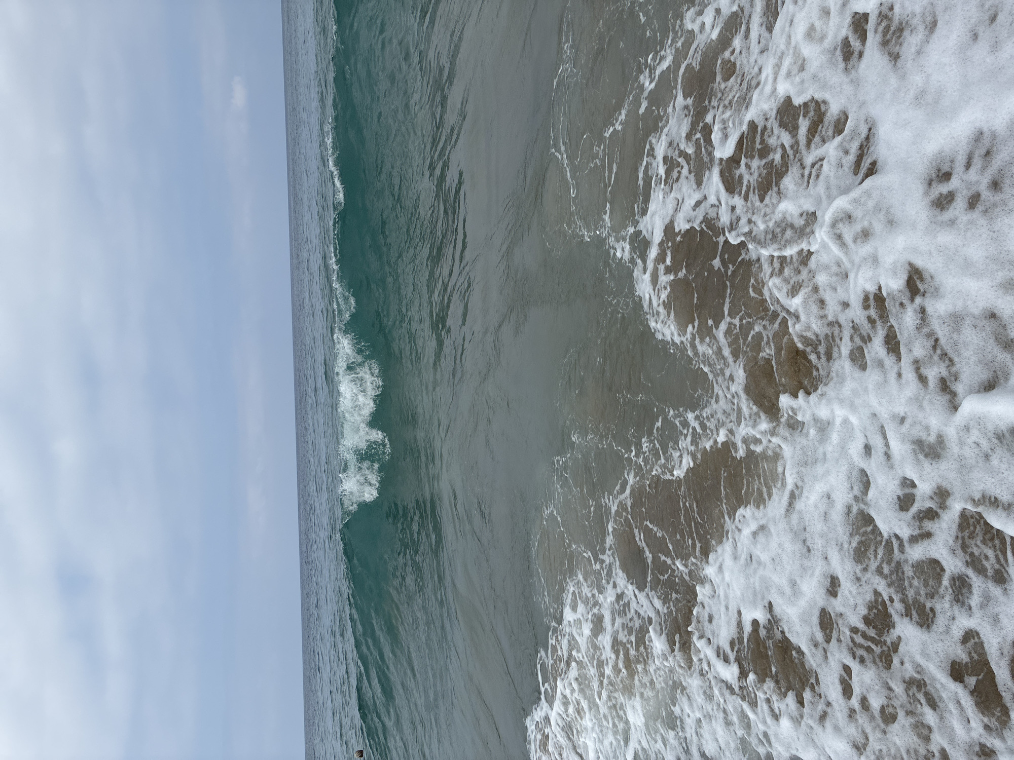 The image depicts a calm ocean with a gentle wave approaching the shore under an overcast sky. The water is a deep teal color, transitioning to a lighter, foamy white as it reaches the sandy beach. In the distance, the horizon is marked by a soft line where the sea meets the sky. There are no discernable landmarks or structures in the image that would indicate a specific location in Alanya, Turkey. The scene is one of natural serenity, with the focus solely on the interplay of water and sand.
