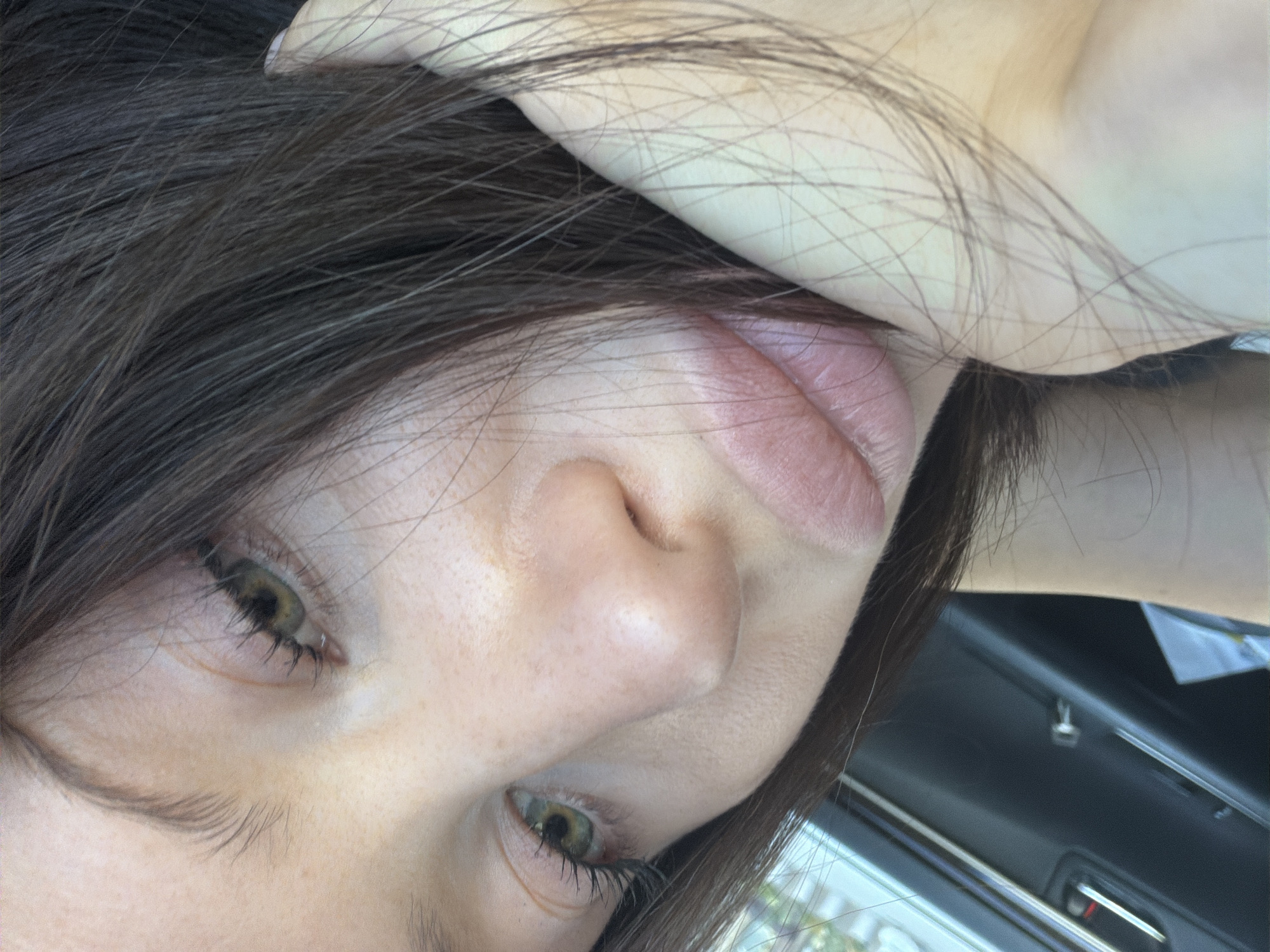 This is a close-up, low-angle, eye-level photograph taken outdoors. The image primarily features the left side of a person's face, including their eye, nose, and lips, with dark hair obscuring parts of their head and face. The person's eye is open and looking towards the right. The skin appears to have a light tone with some freckles visible on the cheek. The lips are light pink and closed.
The background is blurred but suggests an indoor setting, possibly the interior of a vehicle, with visible dark upholstery and what appear to be windows with some light filtering through. There are no discernible objects, text, or specific location cues that would definitively identify the setting beyond being inside a vehicle. The lighting suggests it is daytime, but the time of day or weather conditions are not clear. The overall emotion conveyed is neutral, with no strong indicators of happiness, sadness, or anger. The image is oriented with the top of the person's head towards the left of the frame.