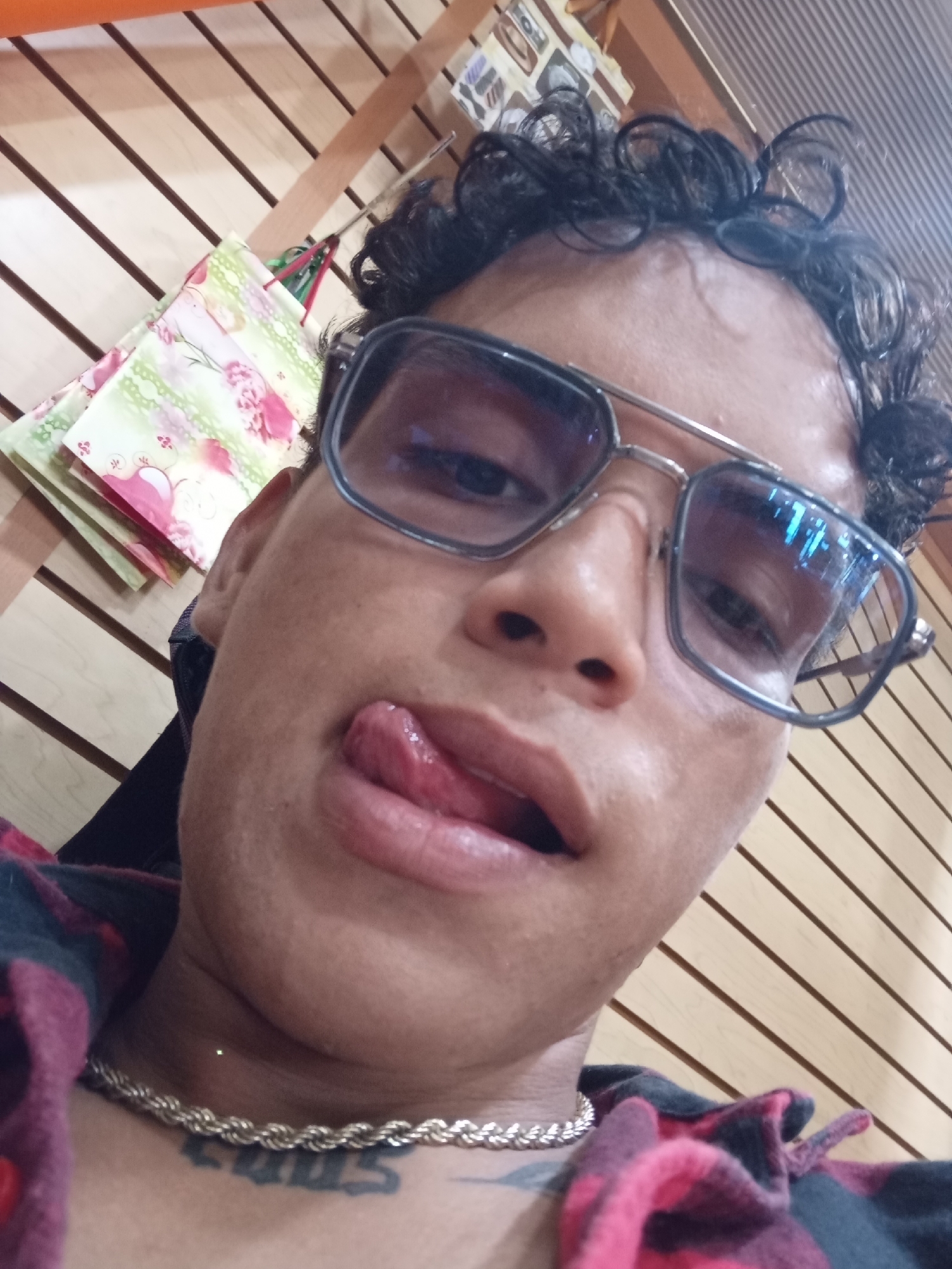 A close-up, low-angle selfie captures an individual indoors, likely in a shop in Juan Griego, Venezuela. The person, an adult with medium skin tone and curly dark hair, is looking directly at the camera with a playful expression, sticking their tongue out. They are wearing large, square-framed sunglasses with dark tinted lenses that reflect blue light on the right side. A silver-colored, thick rope chain necklace adorns their neck, beneath which a dark tattoo with stylized letters, possibly spelling "LOUS" or "LOUIS," is visible. The individual is also wearing a plaid shirt in shades of red and black. The background features a light-colored wooden slat wall, commonly used for retail displays, from which several small decorative bags with floral patterns in light green and pink/red are hanging. The setting suggests a casual, indoor retail environment.