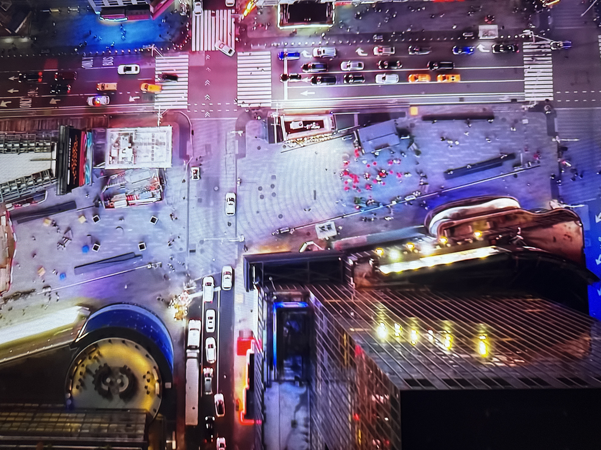 This is an aerial view of a bustling city street scene at night. The image is dominated by the vibrant glow of neon lights reflecting off buildings and pavement. A multi-lane road is visible, with several cars and a bus navigating through the traffic. Pedestrians are scattered across sidewalks and crosswalks, suggesting a lively urban environment.
The composition includes towering buildings with illuminated windows, indicating commercial or residential structures. One building features a large, circular, illuminated structure that appears to be a clock or a decorative element. The street itself is marked with lane dividers and pedestrian crossings.
The overall impression is of a dynamic and active city center at night, filled with light and movement.
The image is located in the city of Burgas, Bulgaria.