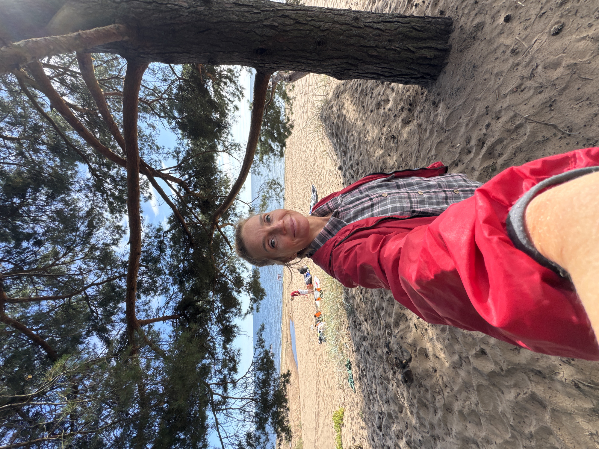 A woman in a red jacket and plaid shirt is taking a selfie on a sandy beach with a pine tree and the Baltic Sea in the background. Several people and beach items are visible in the distance. The scene is located in Zelenaya Roshcha, Russia, a suburban district of Kaliningrad known for its beaches.