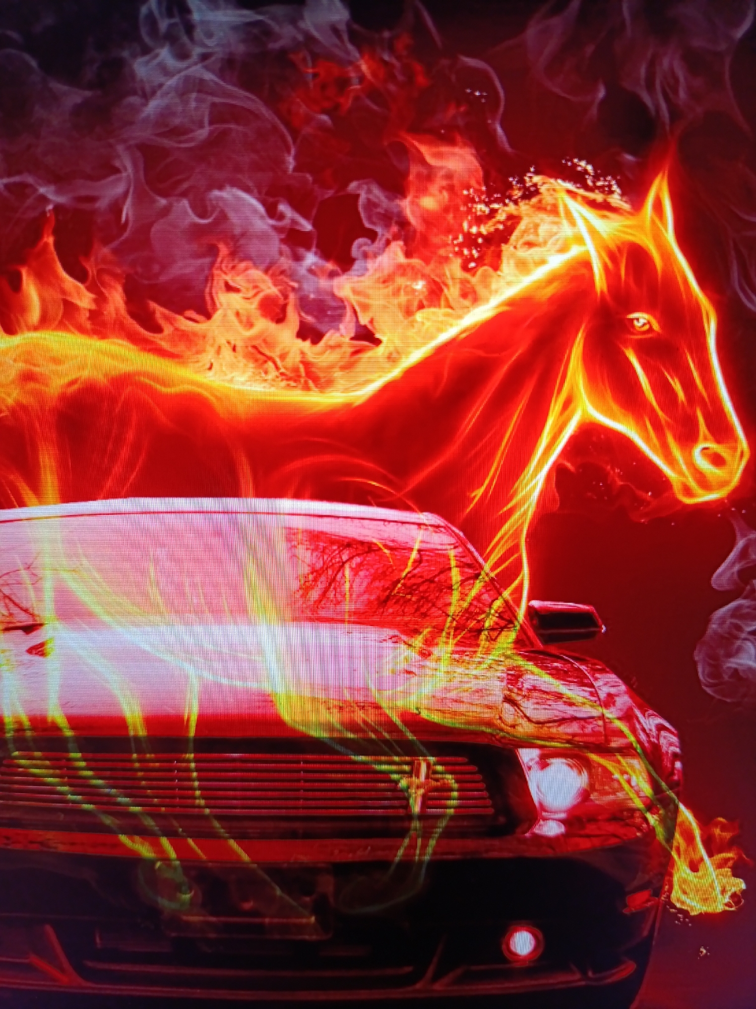 A digitally rendered image depicts a black Ford Mustang with a fiery silhouette of a horse superimposed over it. The horse appears to be made of flames, with its head and neck extending upwards and to the right. The car's hood and grille are visible, with flames also appearing to emanate from the front of the vehicle. The background is a dark, smoky red, with additional wisps of smoke and embers. The image has a high contrast and a vibrant color palette dominated by reds, oranges, and yellows. The composition suggests a powerful and dynamic scene, perhaps symbolizing speed or raw energy. The location is unknown.