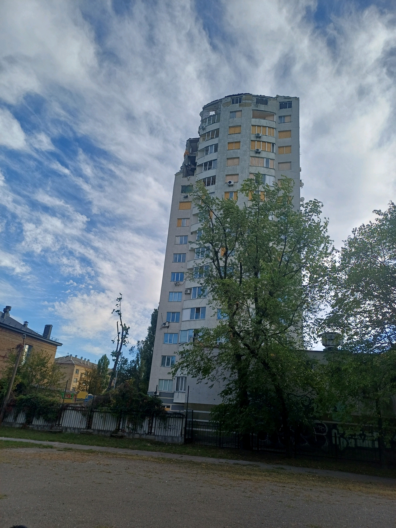 The image captures a damaged multi-story apartment building in Kyiv, Ukraine. The top floors of the building are visibly destroyed, with exposed rebar and missing sections. Many windows on the lower floors are boarded up. The building is surrounded by trees, some of which appear to be damaged as well. In the foreground, there is a dirt path and a fence. The sky is cloudy with streaks of blue. The scene suggests the aftermath of an attack or disaster.