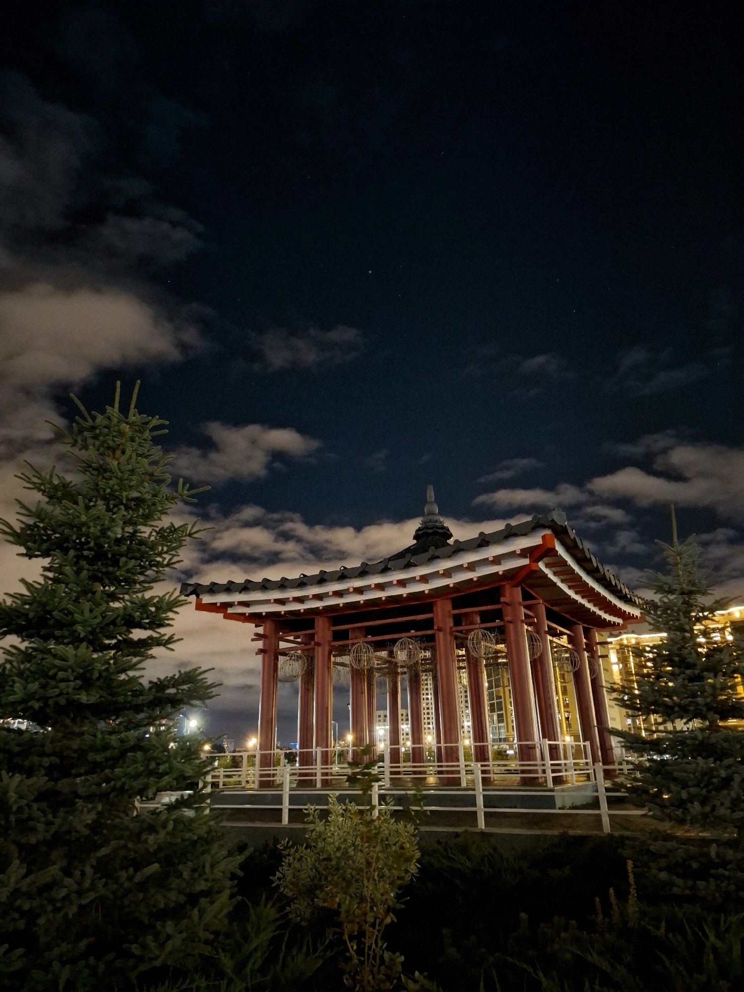 The image shows a night view of a traditional East Asian-style pavilion in the city of Tyumen, Russia, likely located within the "Tyumen Embankment" area. The pavilion has a red painted wooden structure with a multi-tiered, dark tiled roof and white railings. Suspended inside the pavilion are decorative spherical lanterns. To the left and right of the pavilion are dark green evergreen trees, with bushes and grass in the foreground. In the background, tall buildings with illuminated windows are visible, suggesting an urban setting. The sky above is dark blue with scattered white clouds and a few visible stars. The lighting suggests it is nighttime, with the pavilion and background buildings illuminated. There are no people visible in the image.