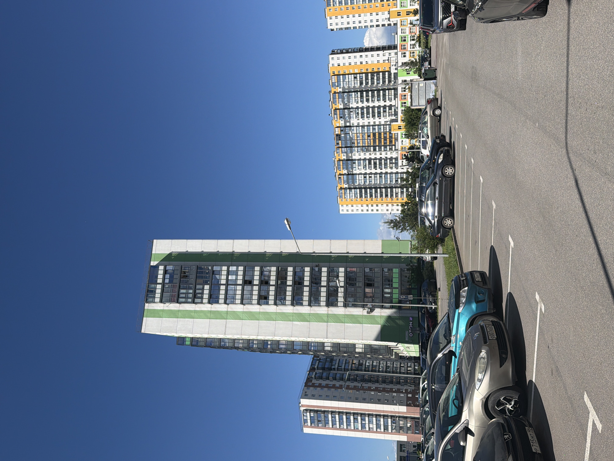 The image shows a bright, sunny day in an urban setting. The sky is a clear, vibrant blue, indicating good weather. The scene is dominated by tall, modern apartment buildings with colorful facades, featuring a mix of white, orange, yellow, green, and brown elements. These buildings are arranged in rows, suggesting a residential complex or a newly developed urban area.
In the foreground and middle ground, a parking lot is visible, filled with several parked cars of various colors, including black, gray, and a striking turquoise blue. The cars are neatly lined up in designated parking spaces marked by white lines on the asphalt. The sun casts long shadows from the cars and buildings, suggesting it is either morning or late afternoon.
One building in the middle ground has a prominent green stripe running horizontally across its facade, with numerous windows. Another building in the background is taller and more densely packed with windows, with a colorful facade that stands out.
There are glimpses of trees and greenery interspersed between the buildings and along the edges of the parking lot, adding a touch of nature to the urban landscape.
There is some visible text on the vehicles, including license plates, but it is difficult to read clearly. There is also some text on one of the buildings that appears to be a logo or signage, but it is also not entirely legible. No people are visible in the image. The overall impression is one of a modern, well-maintained residential area on a clear day.