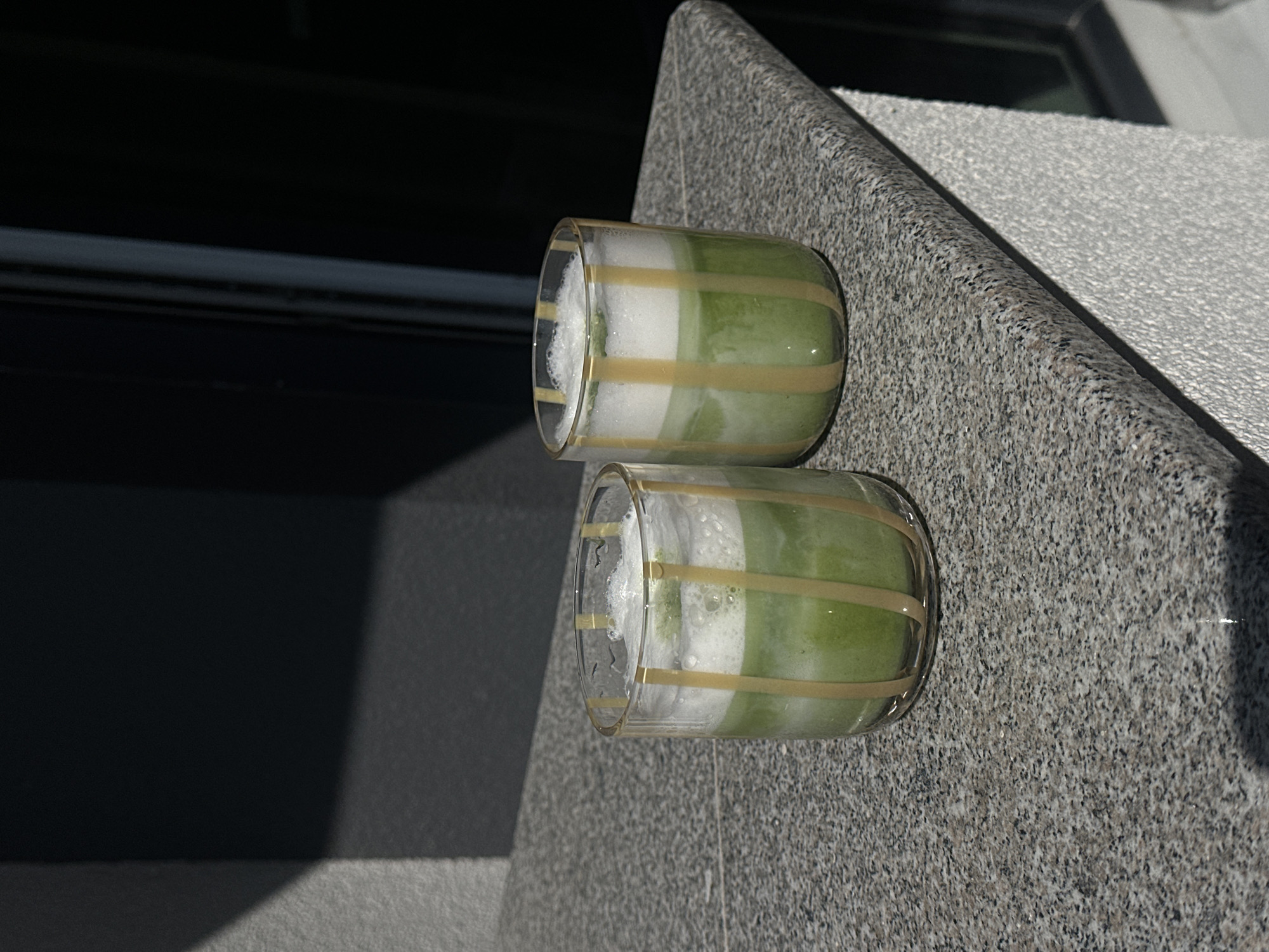 Two striped glasses containing a layered green and white liquid sit on a textured grey surface. The glasses appear to be on the exterior ledge of a building with a dark window behind them. The setting is likely urban given the style of the building and construction material. It's impossible to specify the location as Sofia, Bulgaria, without more identifying context.