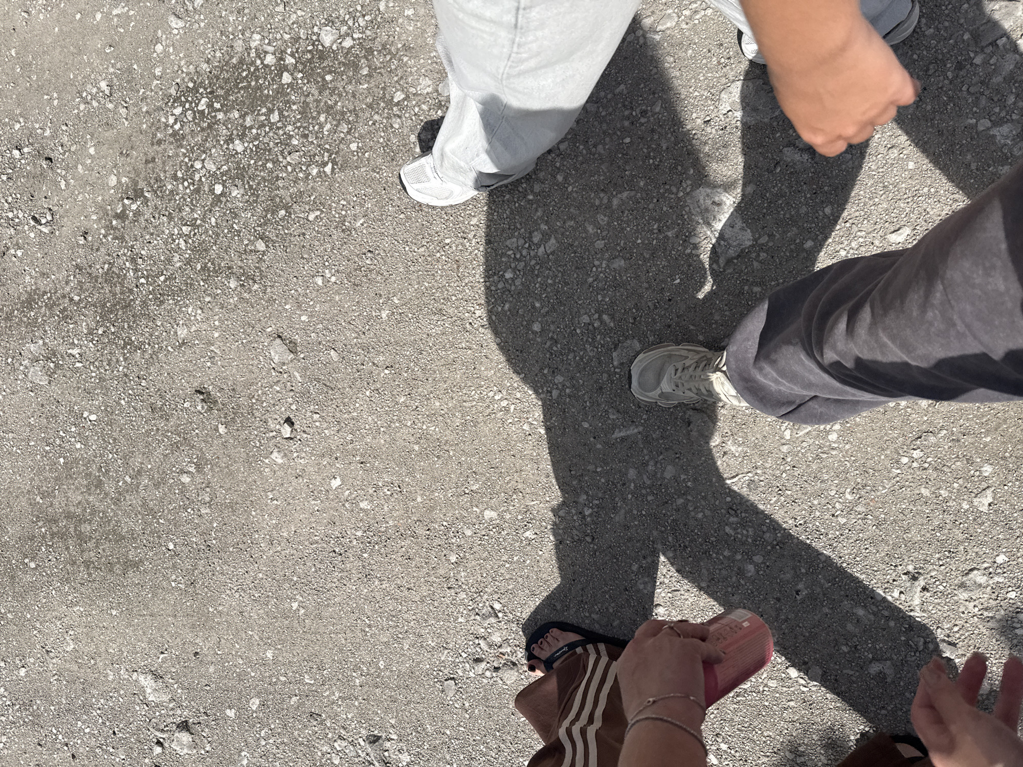 This media file is an overhead shot of several people standing on a gravelly, grey surface. Their legs and feet are visible, wearing various footwear including sneakers and flip-flops. The ground is textured with small stones and pebbles. Shadows are cast across the ground, indicating a sunny time of day. The lighting suggests it is daytime, and the presence of shadows implies fair weather. The scene appears to be outdoors, possibly a pathway or open area. No specific emotions are discernible from the limited view of the subjects. There is no visible text in the media file. The location context is Obukhivka, Ukraine.