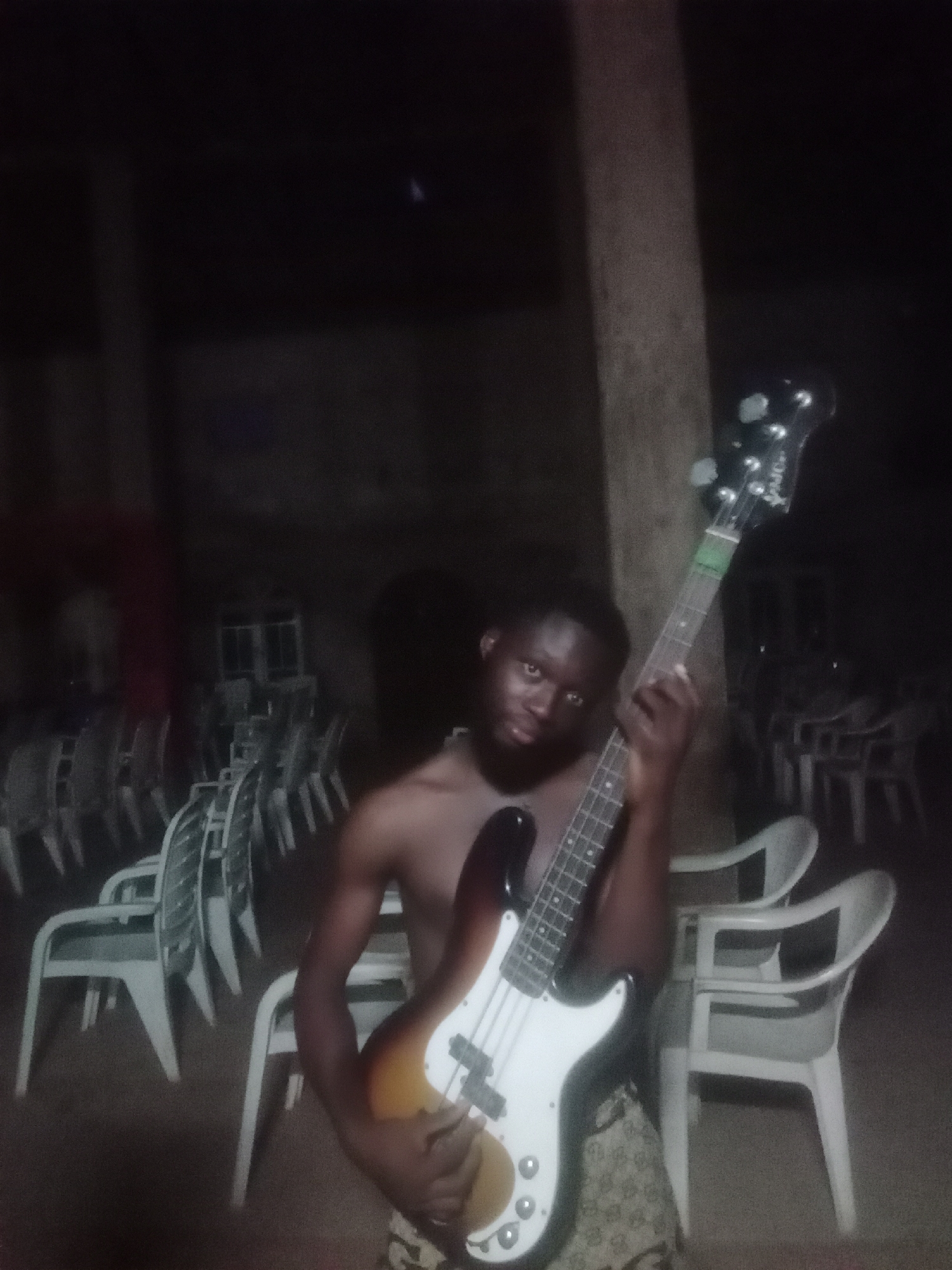 A young, dark-skinned man poses with a bass guitar in a dimly lit, likely outdoor or semi-outdoor setting at night. He is shirtless, wearing patterned shorts, and gazes directly at the camera with a neutral expression.
The man holds a sunburst-finish bass guitar featuring a white pickguard and four strings. Faint text, "Spider" and "MOJO," is visible on the dark headstock. His hands are positioned on the instrument as if ready to play, though he is currently posing.
The background is dark, revealing numerous white plastic chairs, some stacked and others arranged, suggesting a venue such as a hall, event space, or restaurant that is either unoccupied or being prepared/dismantled. A tall, dark pole or tree trunk is visible on the right side of the frame. The low light conditions indicate that the photo was taken at night, with limited illumination accentuating the subject and his guitar. The scene presents a casual moment of the man showcasing his musical instrument in an evening environment, consistent with a setting in Bende, Nigeria.
