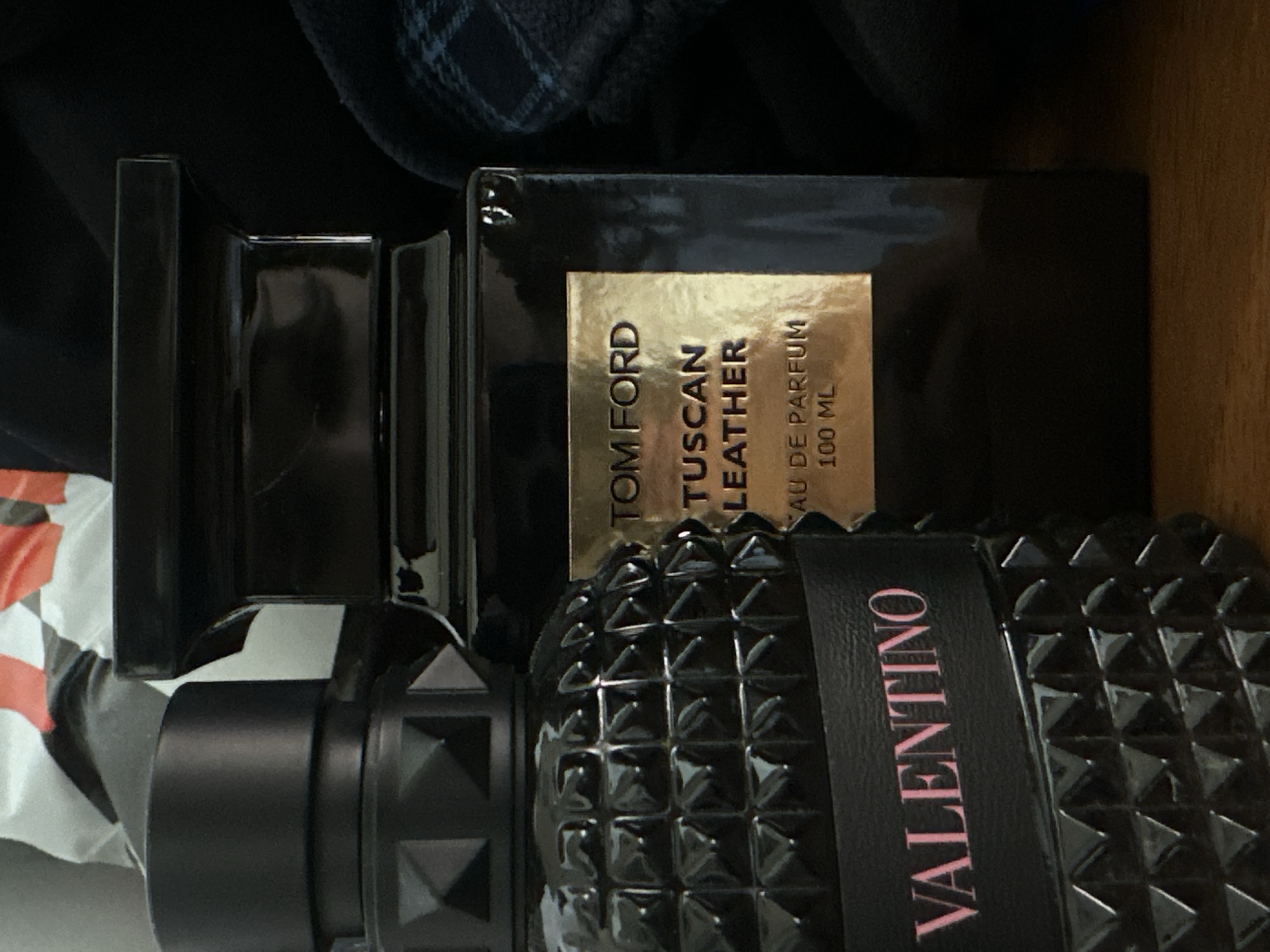 The image displays two perfume bottles on a wooden surface. The bottle on the left is black with a textured surface featuring pyramid-like studs and a band with the word "VALENTINO" in pink lettering. The bottle on the right is also black, with a gold rectangular label that reads "TOM FORD TUSCAN LEATHER EAU DE PARFUM 100 ML." In the background, there are blurred elements of fabric and what appears to be packaging. The scene is lit from above, creating shadows and highlighting the reflective surfaces of the bottles. The location is stated to be Shumen, Bulgaria.