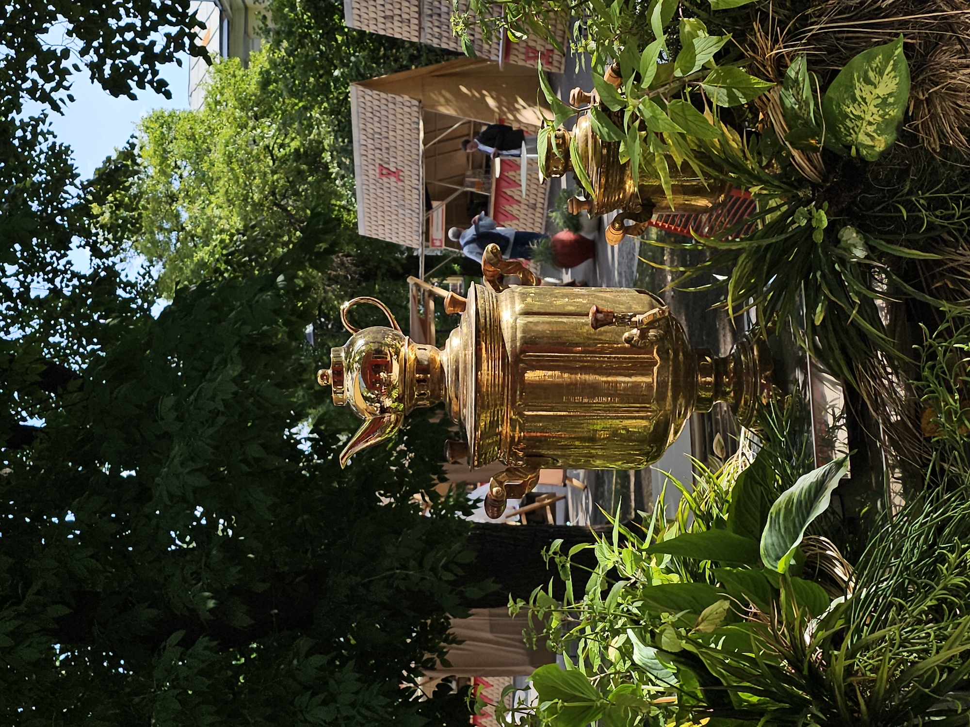 The image depicts a public outdoor setting with lush greenery in the foreground. A large, ornate brass samovar, a traditional Russian tea urn, is prominently displayed in the center. Its polished surface reflects the sunlight and the surrounding foliage. To the right, a smaller, similar brass samovar is partially visible, also adorned with wooden handles. Both samovars are placed on a reflective surface, possibly a tray or a polished base.
In the background, several small, wooden structures with steeply pitched roofs, resembling market stalls or kiosks, are set up. These structures are thatched with wood shingles and feature red detailing. One stall has a counter with what appears to be beverages or food items. Two men are visible near the stalls: one, wearing a blue shirt and a backpack, is standing and looking towards the stalls, while another, in a dark jacket, is standing by a counter, possibly interacting with someone off-camera. Beyond the stalls, mature trees with dense green foliage create a canopy, and parts of buildings with light-colored facades and tiled roofs are visible. The sky is bright blue with some clouds.
While the specific location within Moscow, Russia, cannot be determined solely from the visual information, the presence of samovars, the architectural style of the stalls, and the overall atmosphere suggest a cultural event, market, or festival likely taking place in a park or public square in Moscow. The scene is bathed in natural daylight, indicating it was taken during the daytime.