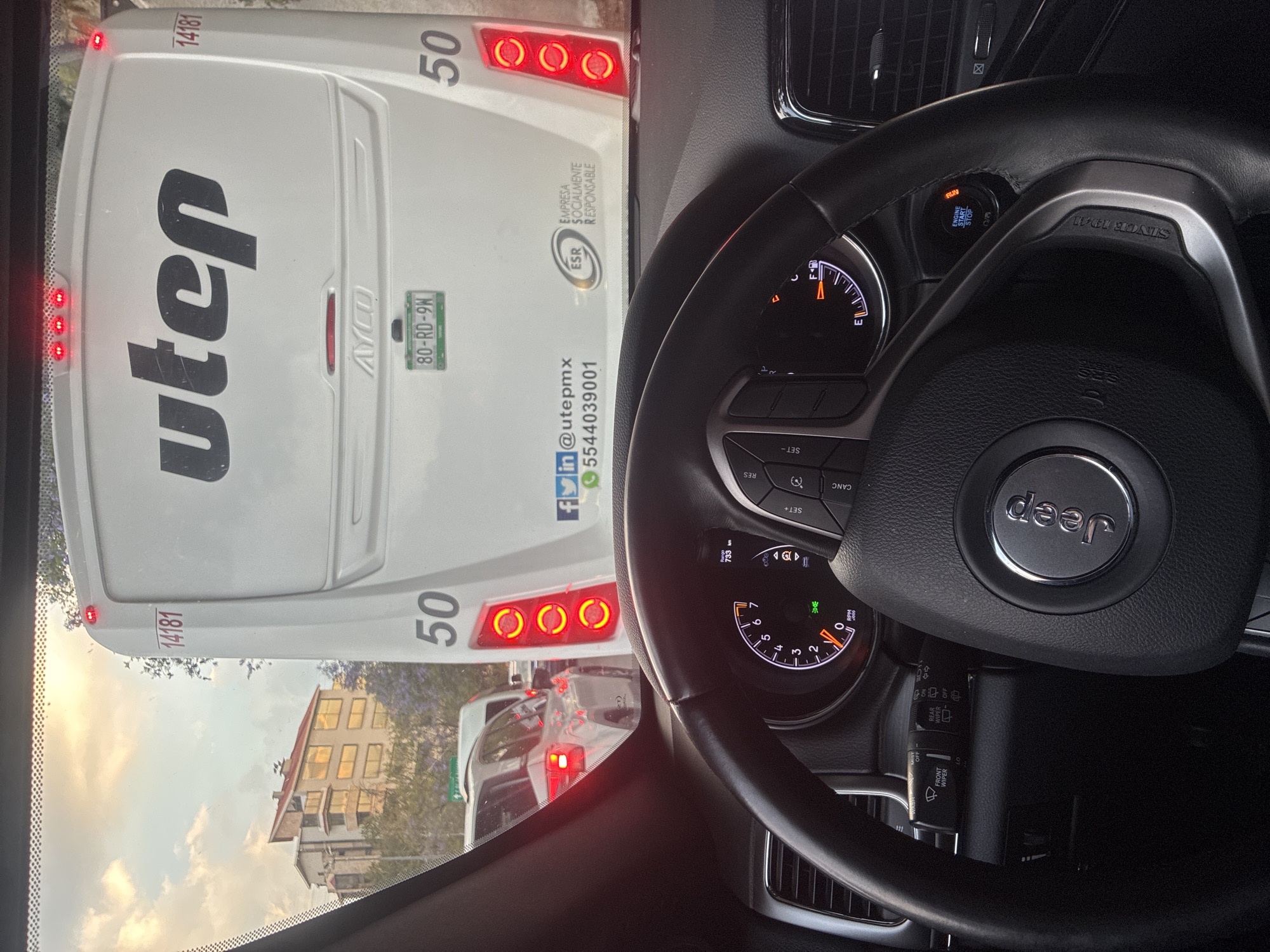The view from the driver's seat of a Jeep shows a UTEP bus directly in front. The bus, numbered 50 and marked "14181," displays the license plate "80-RD-9N," social media contacts, and the logo of a socially responsible company. A building is visible through the windshield. The scene appears to be set in Mexico City, Mexico, given the UTEP bus which is based in the area. The Jeep's dashboard is visible, and the car appears to be running.