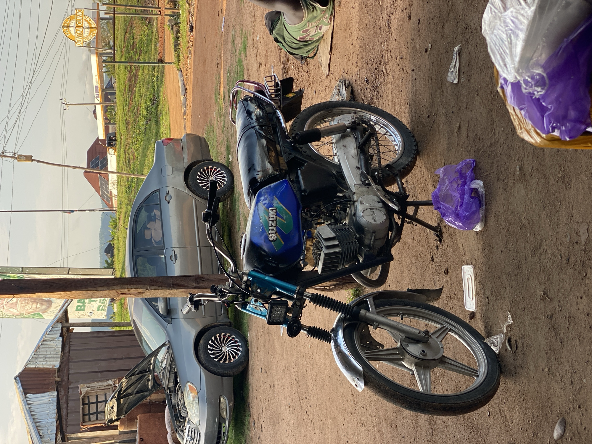 The image depicts an outdoor scene in Jalingo, Nigeria, with a dirt ground surface. In the foreground, a blue and black Suzuki motorcycle is parked, leaning on its stand. It has a silver front fender and spoked wheels, with a black seat and handlebars. To the right of the motorcycle, a person is lying down on a piece of cardboard, wearing a green tank top and dark shorts.
Behind the motorcycle, a grey sedan is parked with its hood open, revealing the engine. The car has custom wheels with black and silver spokes. To the left of the car is a small, rustic structure with a corrugated metal roof and wooden walls. In the background, there is a dental clinic sign and a gas station. The sky is overcast, and the overall environment suggests a roadside or commercial area. There are several plastic bags scattered on the ground.