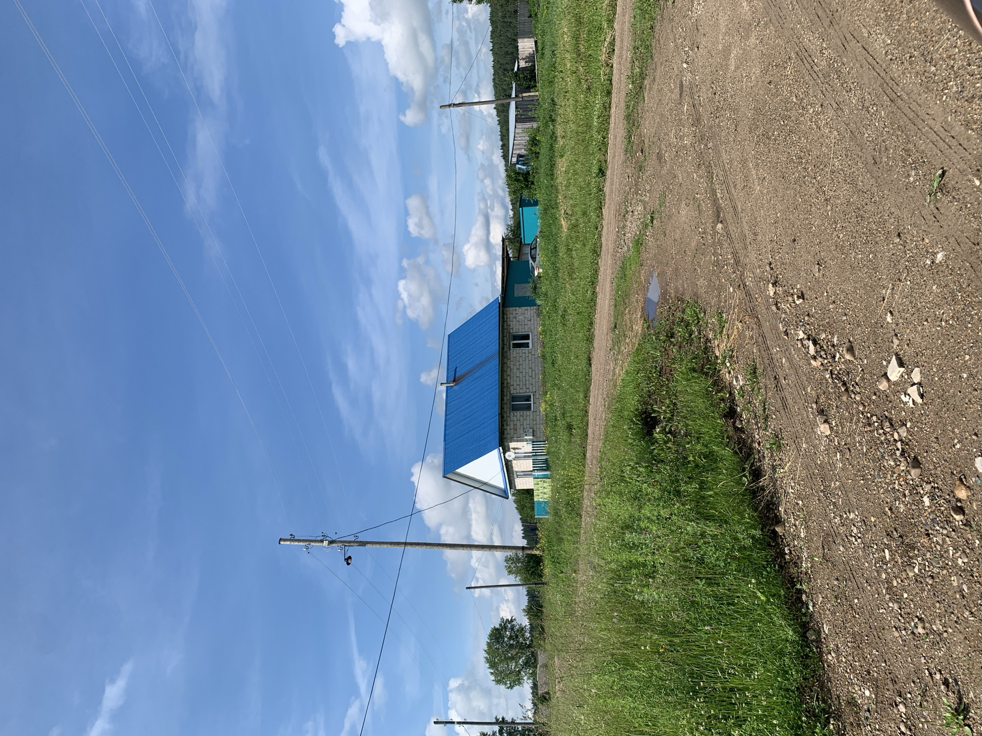 The image depicts a rural scene in Barsai, Russia, under a bright blue sky with scattered white clouds. A dirt road, with visible tire tracks and scattered pebbles, leads into the frame from the foreground, curving slightly to the right. To the left of the road, tall green grass and other vegetation grow.
In the middle ground, a light-colored brick house with a distinctive bright blue, sloped roof stands prominently. The house has a single window visible on its side and a small green fence in front of it. Adjacent to the house, there is a teal-colored shed or outbuilding. Further to the right, another outbuilding with a wooden exterior and a metal roof is visible, along with a wooden fence and a dense line of trees forming the background.
Several utility poles with wires stretch across the sky, indicating the presence of electricity and possibly telephone lines. One pole is located to the left of the house and another to the right, closer to the background. A parked white car is partially visible near the teal outbuilding. The overall environment is natural and appears to be a residential area in the countryside.