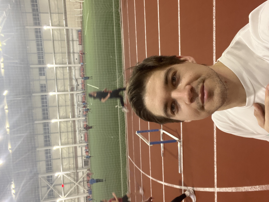 A man in a white shirt is taking a selfie indoors. He's on a red running track with white lane markers. In the background, there's a green soccer field behind a net. Several people are on the field. A hurdle is also visible on the track. The building has a high ceiling with lights and beams.