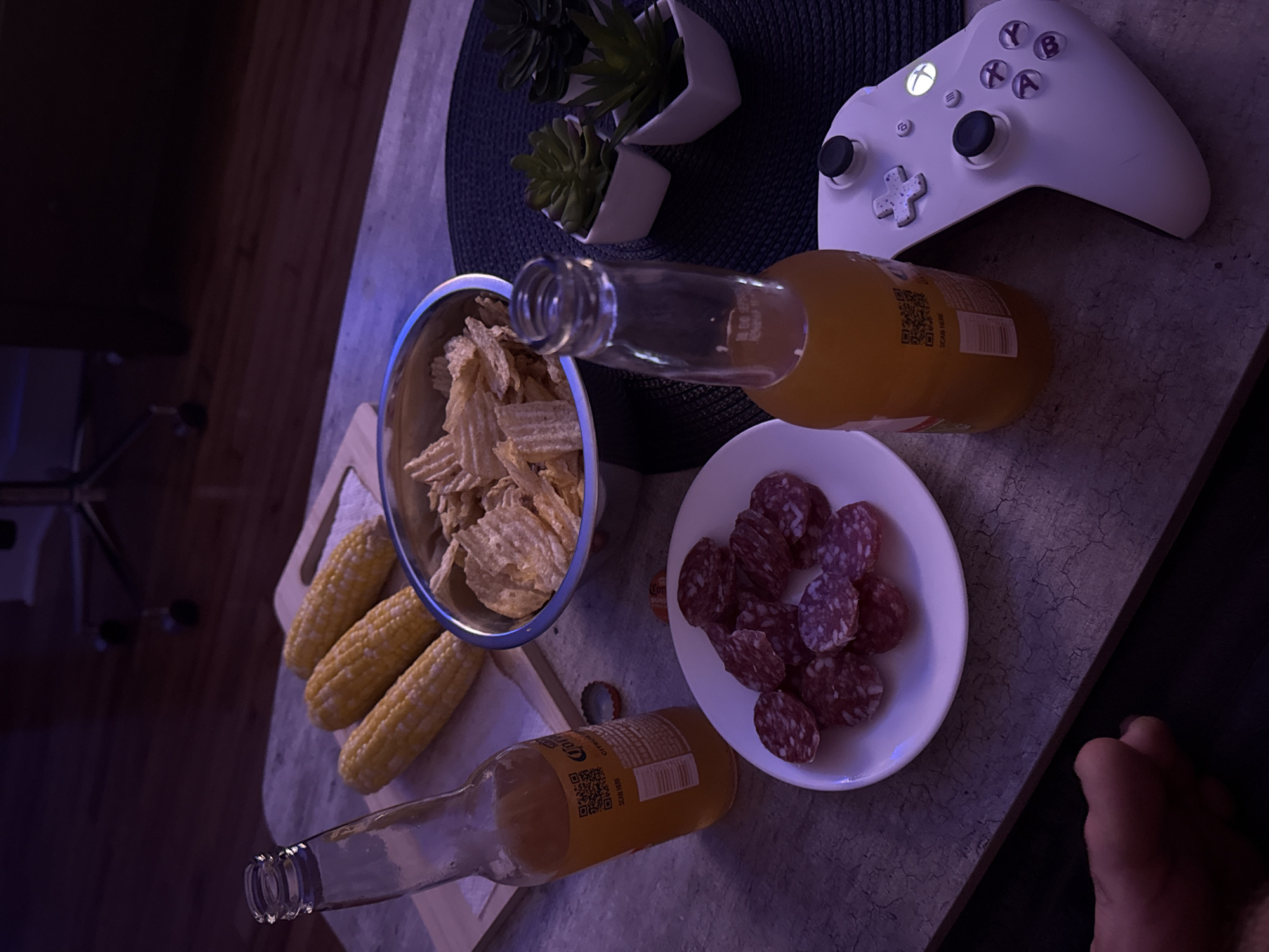 This is a still-life photograph taken indoors, likely in a living room or entertainment area, suggesting a casual setting. The scene is lit with dim, atmospheric lighting, creating a moody ambiance.
The primary subjects are food and drink items arranged on a textured, grey coffee table. In the foreground, a white plate holds several slices of cured sausage, appearing to be salami. To the right, a white Xbox controller is prominently displayed, its power button illuminated. Adjacent to the controller, a glass bottle with a yellow liquid, possibly beer or soda, stands upright.
Further back on the table, a metal bowl is filled with golden-brown potato chips. Three ears of corn, husked and yellow, are placed on a wooden cutting board behind the chips. Another identical glass bottle is situated to the left of the corn. The table itself has a rough, concrete-like texture.
In the background, the scene transitions to darker tones, revealing wooden flooring and the lower portion of a piece of furniture with wheels, possibly a desk chair. Three small potted plants with green succulent-like foliage in white square pots are arranged on a dark placemat to the right of the table.
There are no individuals directly visible in the frame, though a hand with fingers is partially in the bottom left corner of the image, suggesting someone is off-camera, perhaps holding or interacting with the scene. The overall impression is one of a relaxed evening, potentially involving gaming or watching something. The specific location within the city of New York, United States of America, cannot be determined from the visual information provided.