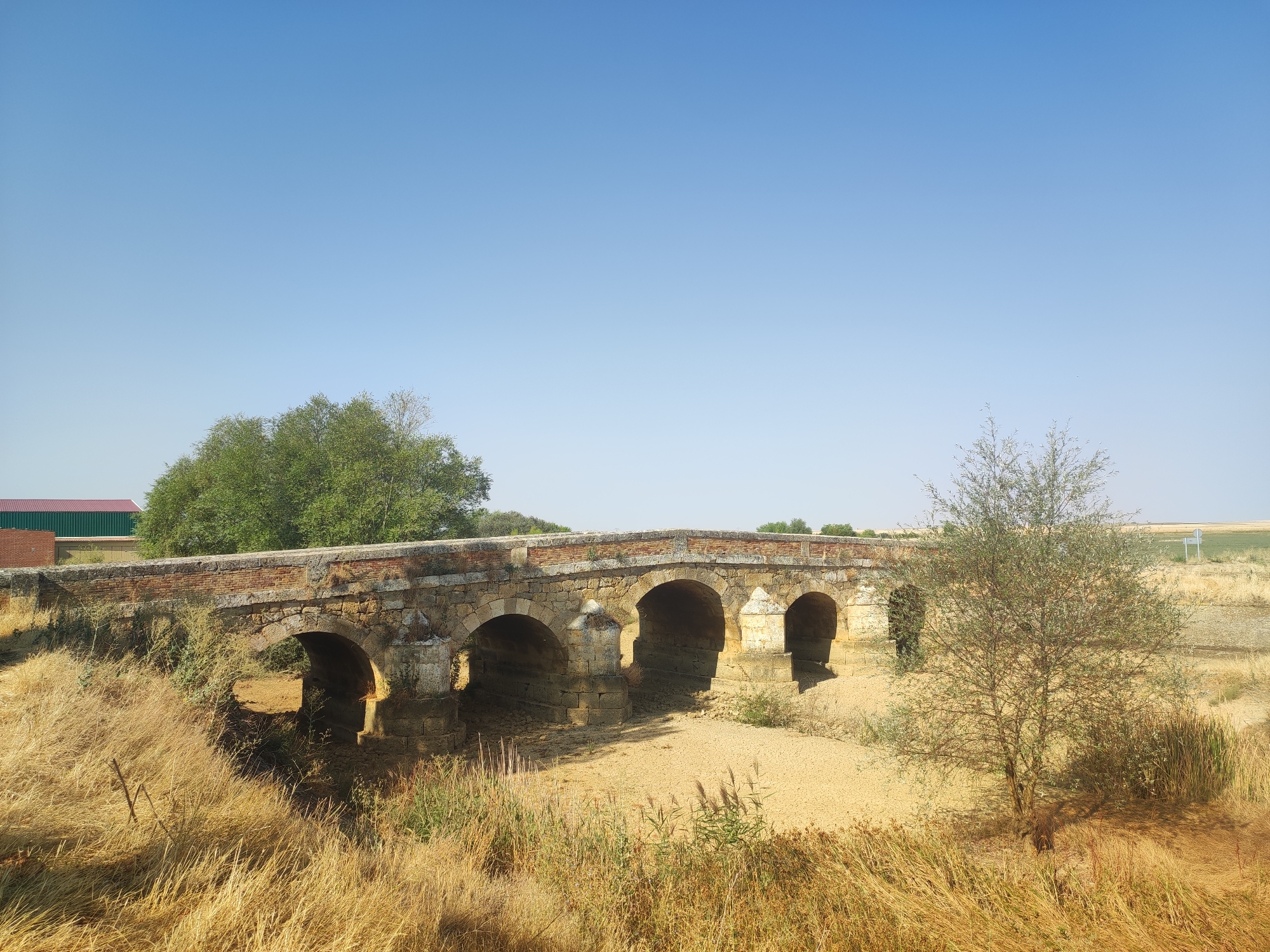The image depicts a stone bridge with multiple arches spanning a dry riverbed. The bridge is made of weathered stone blocks and brickwork, with vegetation growing along its edges. The surrounding landscape is arid, with dry grass and sparse trees. In the background, there are some buildings and agricultural fields under a clear blue sky. The scene is set in Autillo de Campos, Spain, a location known for its historical bridges and agricultural significance. The overall impression is one of timelessness and natural beauty, with a touch of rustic charm.