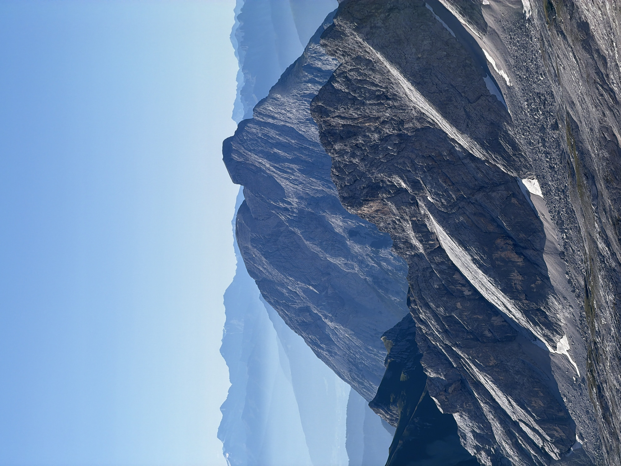 The image depicts a mountainous landscape with several layers of peaks receding into the distance under a clear blue sky. The foreground features rugged, rocky slopes with patches of snow. The middle ground is dominated by a large, imposing mountain with a distinct peak and textured rock faces. In the background, progressively lighter and more mist-covered mountains create a sense of depth. The scene is devoid of any people or man-made structures. This is a view of the Alps, likely from a high vantage point in or near Garmisch-Partenkirchen, Germany, given the characteristic mountain formations.