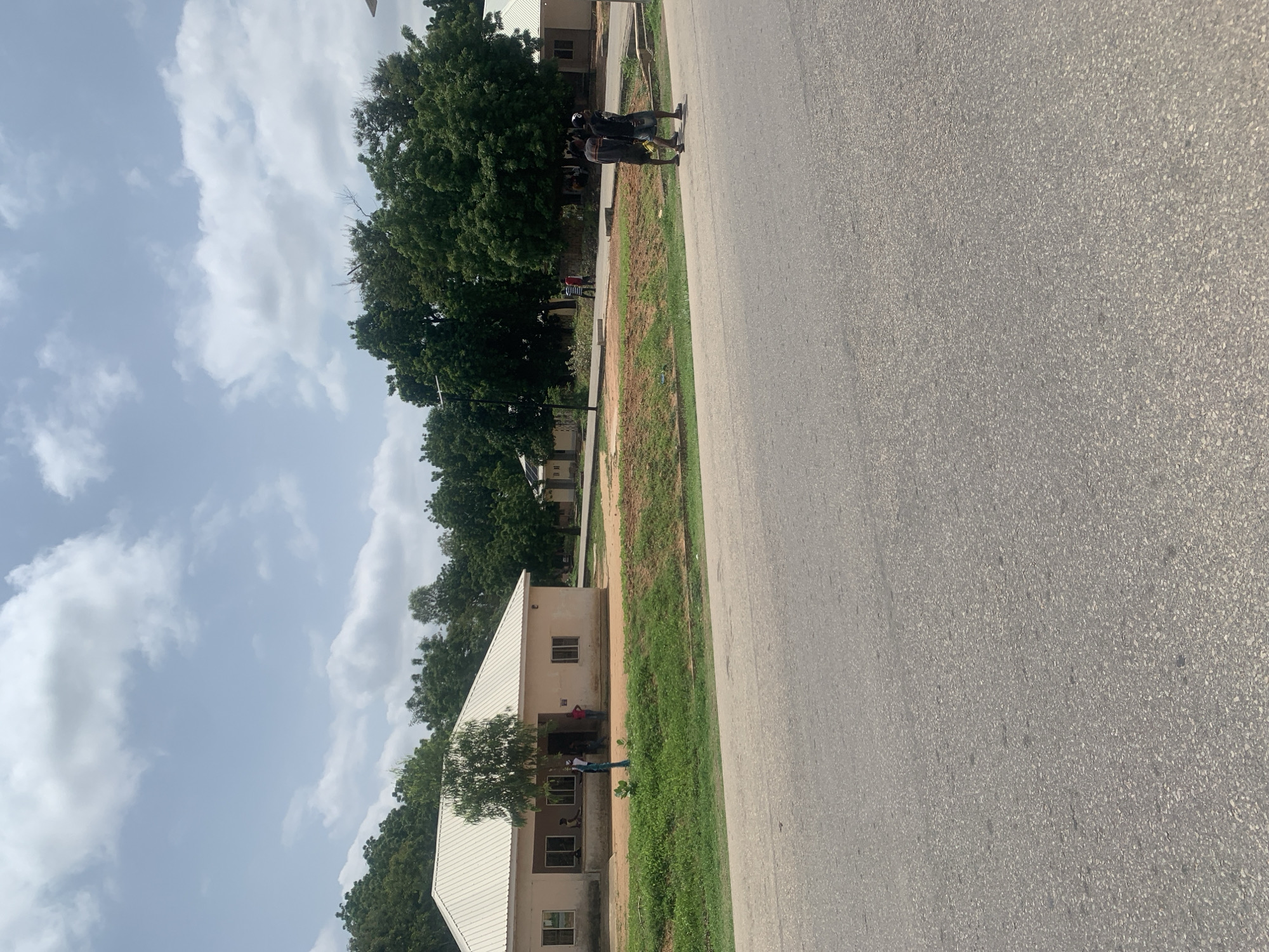 The image depicts a street scene in Jalingo, Nigeria, likely on a university campus or similar institutional setting. The foreground is dominated by a paved road. In the middle ground, a single-story building with a light-colored, sloped roof is visible, flanked by greenery and trees. Several individuals are seen near the building, some appearing to interact or stand by the entrance. To the right of this building, another similar structure is partially visible. Further in the background, a dense line of lush green trees forms a natural boundary. The sky above is bright with scattered white clouds, suggesting daytime. Two individuals are walking on the paved area to the right of the main building, facing away from the viewer. Another group of individuals can be seen near the entrance of the first building. The overall impression is of a public outdoor space in a warm climate.