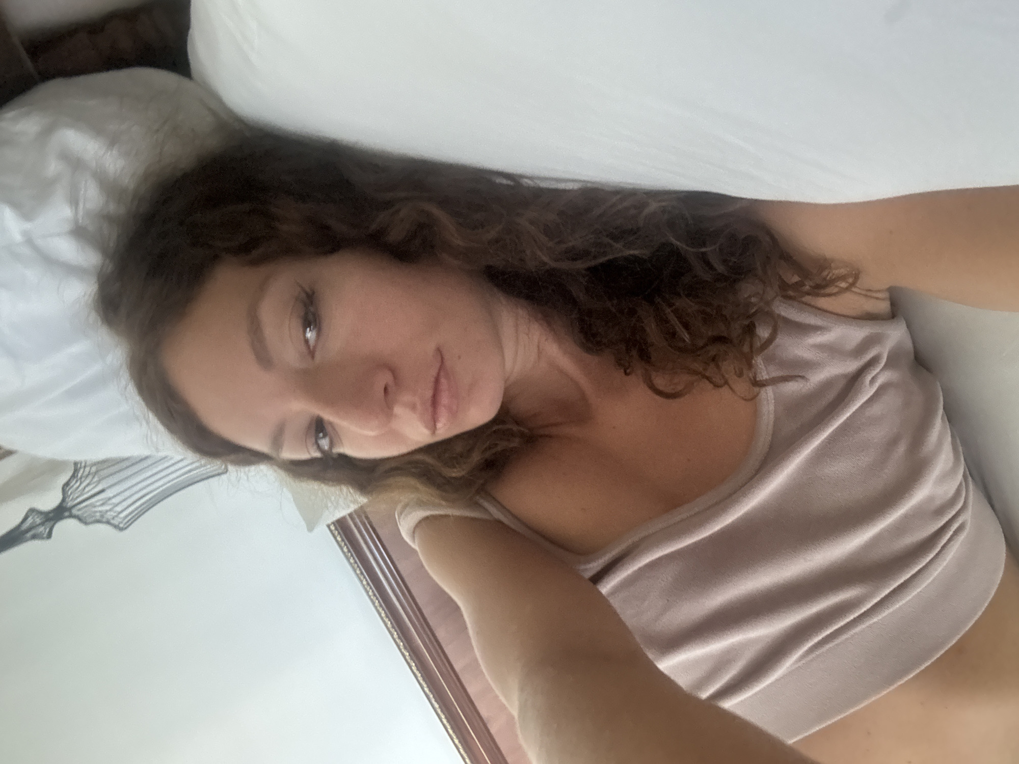 A woman with curly brown hair lies on her left side on a white bed. She is wearing a light-colored top. Her head rests on a white pillow. The lighting is bright and appears to be from a window. The location is not discernible from the image.