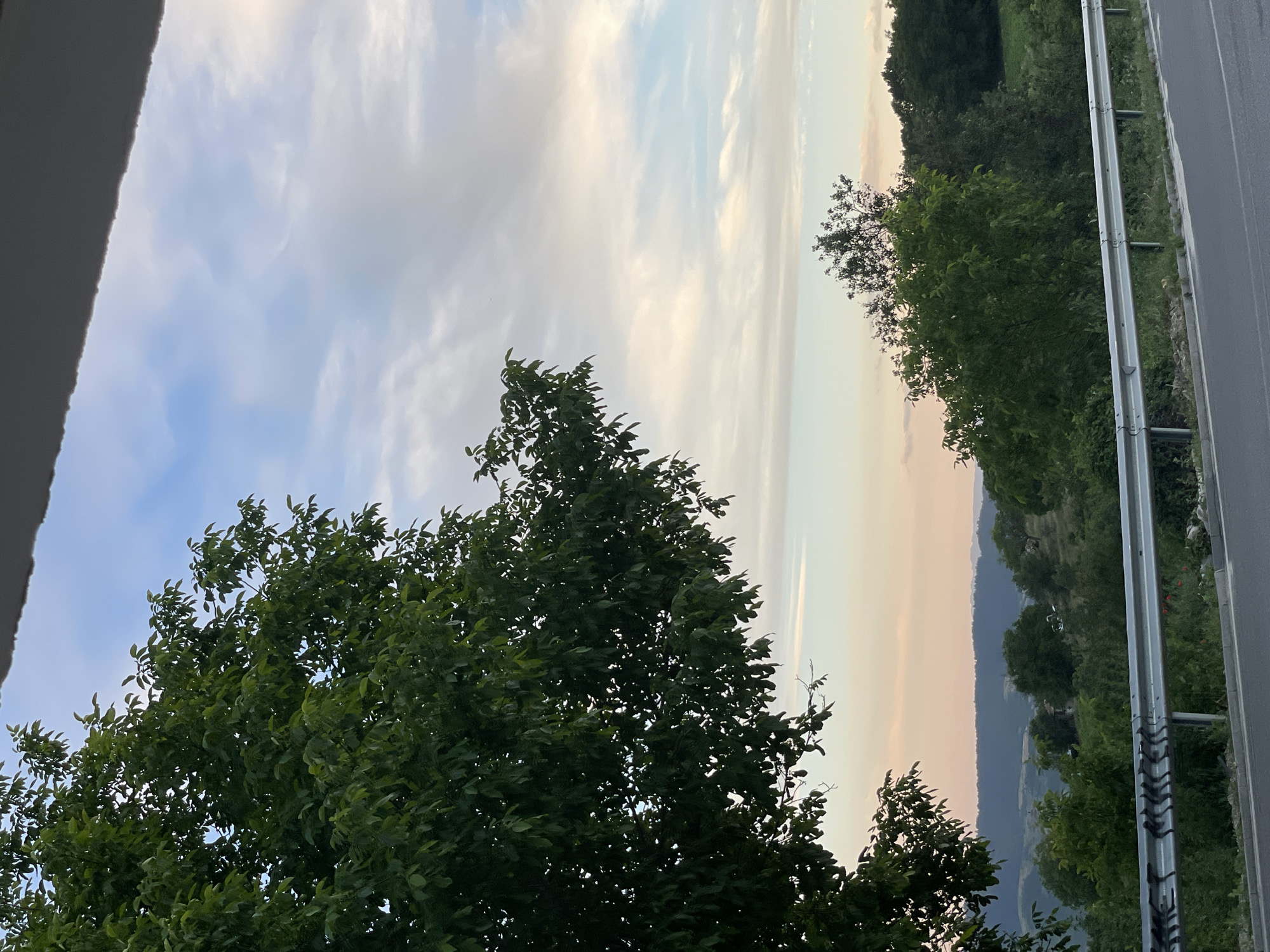 The image depicts a scenic view of a rolling landscape under a sky with scattered clouds, suggestive of late afternoon or early evening. A prominent tree with dark green foliage occupies the left side of the frame, its branches extending into the upper portion of the image. Below the tree, a metal guardrail runs horizontally along the edge of an asphalt road, separating it from a vegetated embankment. In the background, hills and valleys covered in lush greenery stretch towards the horizon, with hints of buildings or structures visible amidst the trees. The sky transitions from pale blue near the horizon to a lighter, cloud-filled expanse above.
The specific location is identified as Valkosel, Bulgaria, known for its picturesque natural surroundings. The scene captures a serene moment, characteristic of rural Bulgarian scenery. There are no people or animals visible in the image, nor any discernable actions or interactions taking place. The focus is on the natural environment and the road infrastructure.