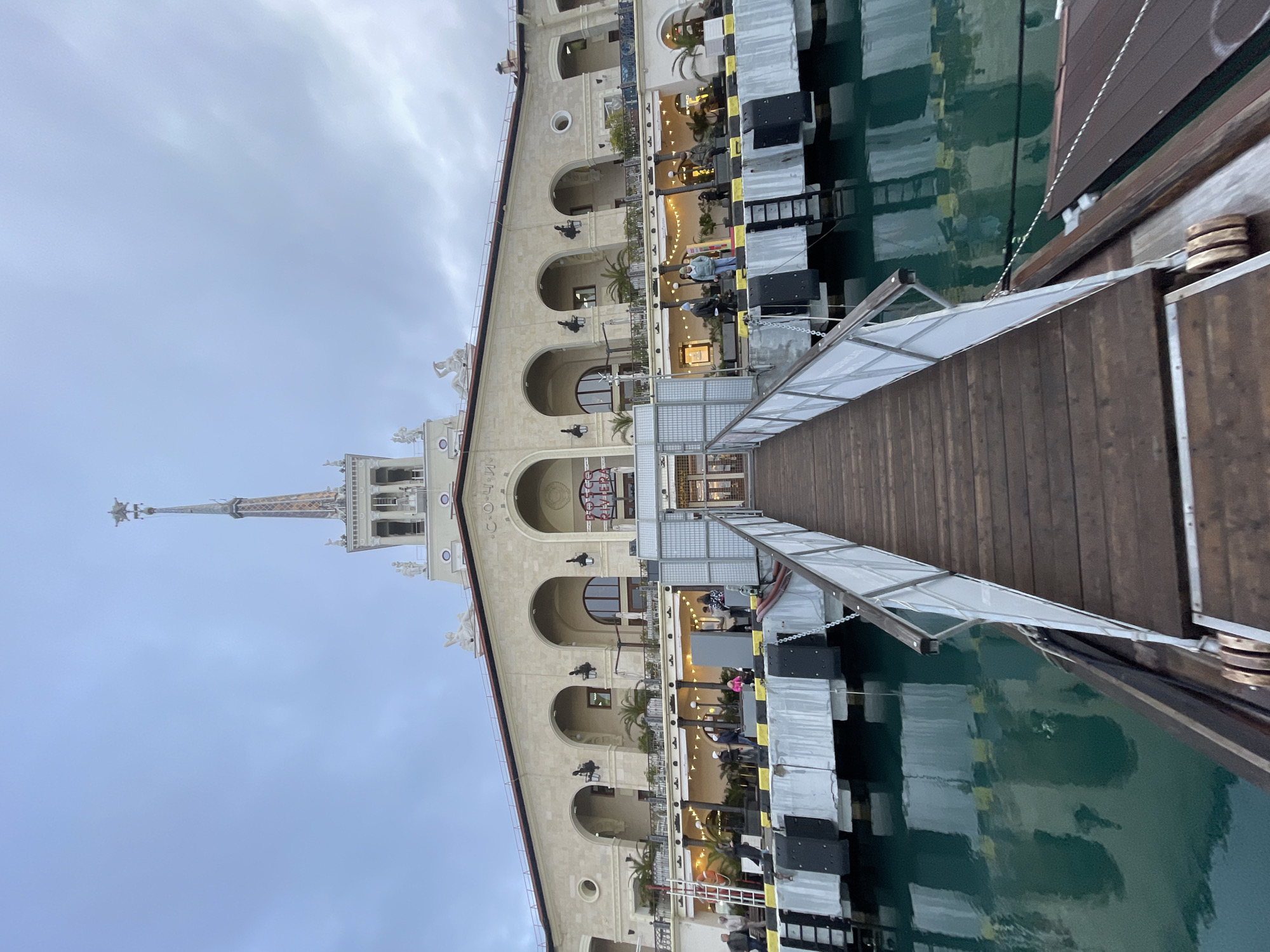 An upward-angled, eye-level photograph captures the Sochi Sea Port Terminal building from the waterfront. In the immediate foreground, a dark wooden gangway with white side railings extends from the bottom center of the frame towards the terminal. To the left and below the gangway, calm, dark turquoise water reflects parts of the pier structure.
The midground features a concrete pier or dock structure, parallel to the main building. This pier has dark grey/black fenders and yellow-and-black striped bumpers. A metal ladder is attached to the pier on the left. Several individuals are visible on the pier; some stand near the railing, others closer to the building.
The central subject is the multi-story, light-colored architectural structure of the Sochi Sea Port Terminal, characterized by neoclassical design elements. The facade features a series of prominent arches on the upper and lower levels, supported by columns or pilasters. Balconies or terraces are present on the lower level, adorned with string lights, potted palm trees, and other foliage, suggesting active commercial spaces. Individuals are also visible on these terraces.
A tall, slender spire rises centrally from the building's roof, topped with a five-pointed star. The upper roofline features classical statues, including reclining figures. The word "СОЧИ" (Sochi) is visible on the building's upper facade to the right of the spire, with "ROSA RIVIERA" displayed in red neon below. The sky overhead is overcast with a uniform light grey cloud cover.