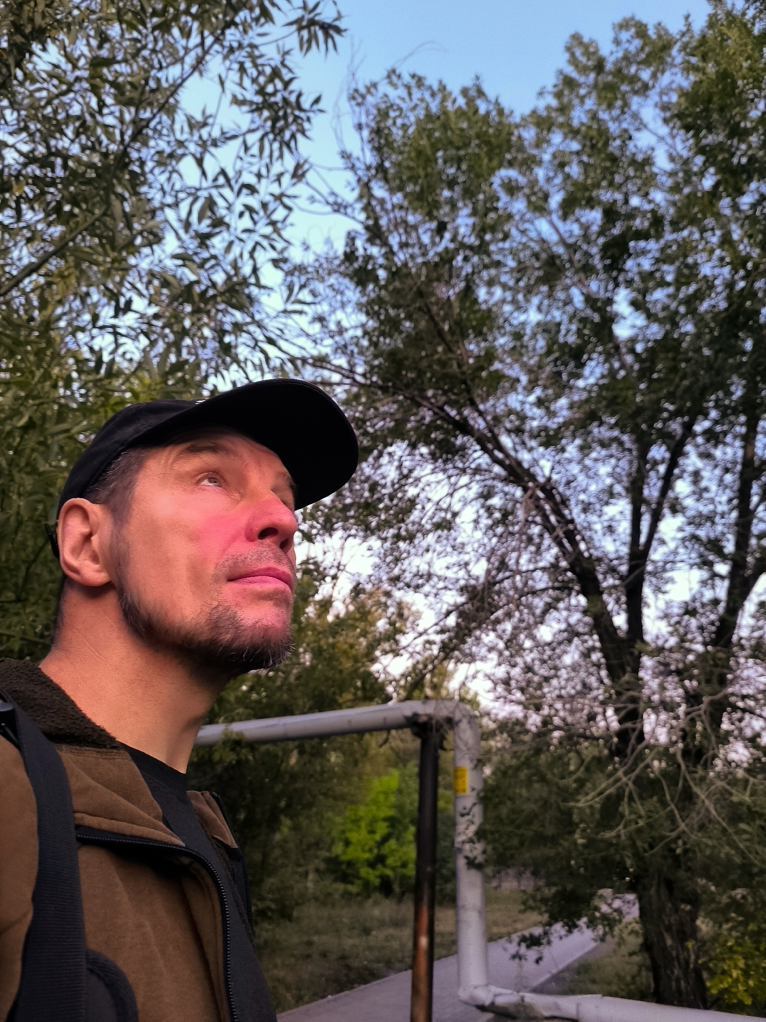 A man wearing a black baseball cap and a brown zip-up jacket looks up and to the right. His face is illuminated by warm light, casting a pinkish hue on his cheek. He has a short beard and is looking upwards. The background consists of trees with green leaves and a pale blue sky. In the mid-ground, a section of piping is visible. The scene appears to be outdoors, possibly in a park or urban green space. No specific identifying features of Qarağandı city administration are apparent in the image.