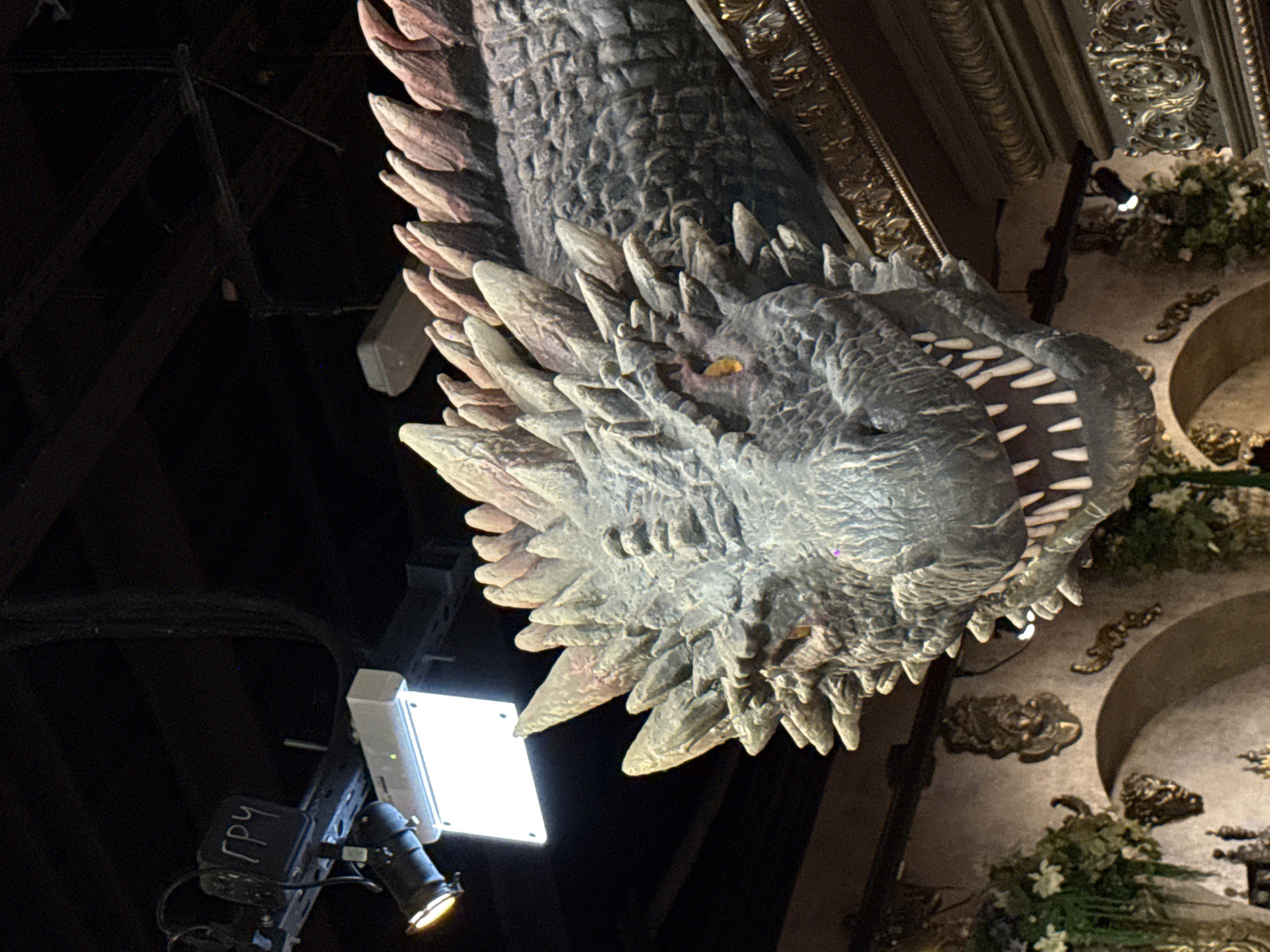 The image shows a large, hanging dragon head, likely a decoration or exhibit. The dragon is gray, with numerous spikes and teeth, and its mouth is open. The lighting is dramatic, with a spotlight illuminating the dragon from below. In the background, architectural details of the building are visible, including ornate decorations and moldings. A lighting rig is also visible in the lower left corner. The image was taken inside the Central Children's Store in Moscow, Russia.