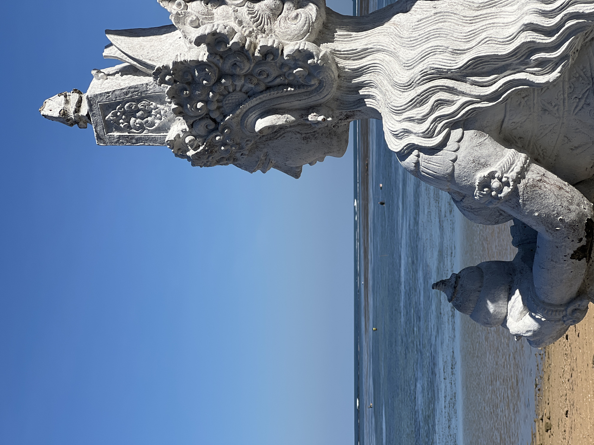 The image shows a weathered, white stone statue, possibly a Balinese deity, positioned on a sandy beach in Denpasar, Indonesia. The statue's head and upper torso are visible against a clear blue sky. The statue's features include intricate carvings and ornate details on its headpiece and hair. The background consists of a calm ocean with a visible horizon line. The sandy beach extends towards the foreground, with the lower portion of the statue's arm and hand holding an object resting on the sand.