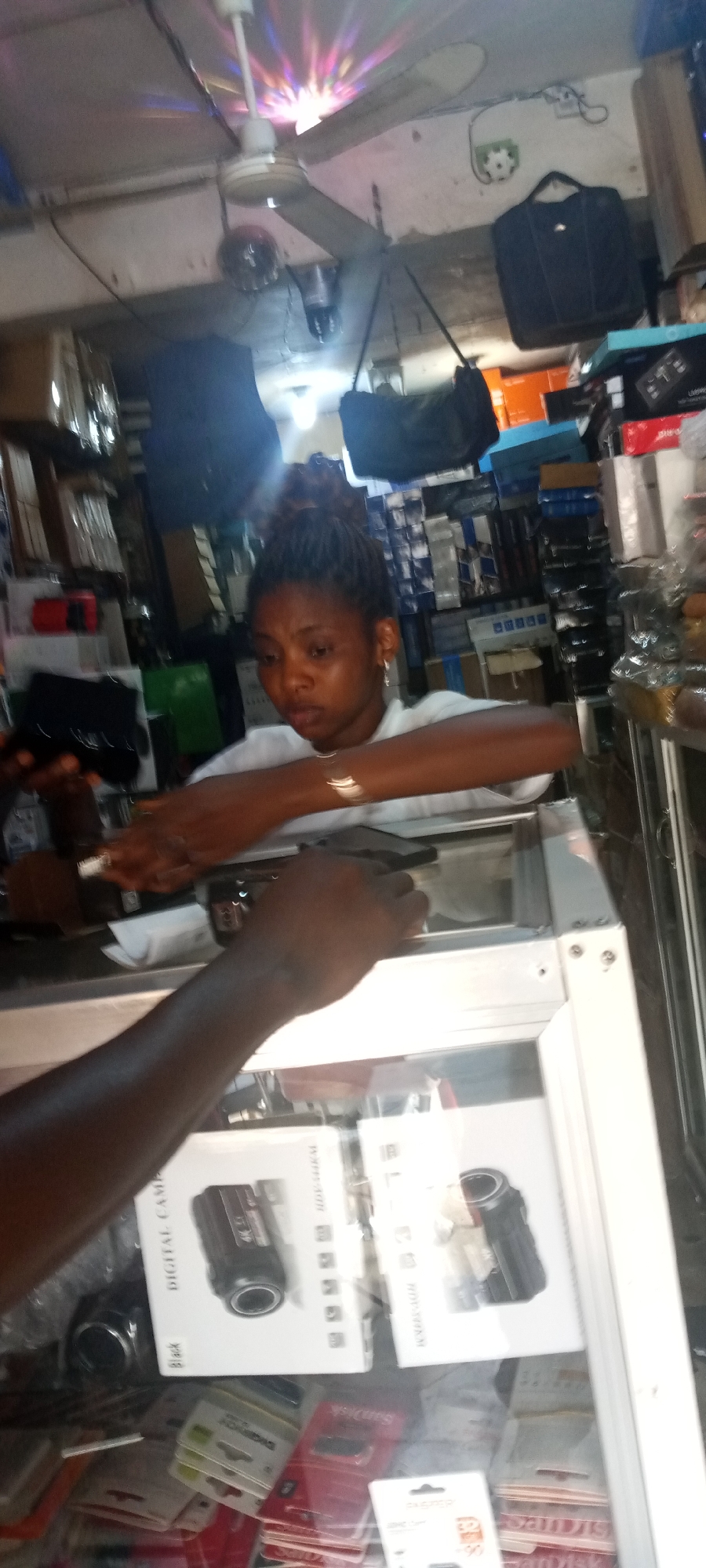 The image shows a woman with braided hair in a bun, wearing a white shirt and jewelry, standing behind a glass display case. She is interacting with a customer, whose dark-skinned arm and hand are visible in the foreground holding a black device. The display case contains items such as digital cameras in their packaging and SD cards. In the background, there are shelves stacked with various items and bags hanging from the ceiling. A ceiling fan with a disco ball is visible above, casting colorful light patterns. The location is a shop in the city of Unknown, Unknown.