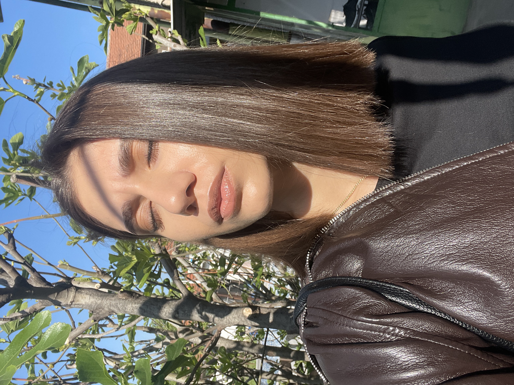 A medium shot depicts a young woman with fair skin, brown hair, and closed eyes in an outdoor setting. She wears a black shirt and a brown leather jacket with a visible zipper. A gold chain necklace is also visible. She is positioned near a tree with green leaves and branches, partially obscuring the background, which includes a building with a red tile roof and a blue sky. The scene is lit by natural sunlight, casting shadows on her face and hair. The location appears to be in Sofia, Bulgaria.