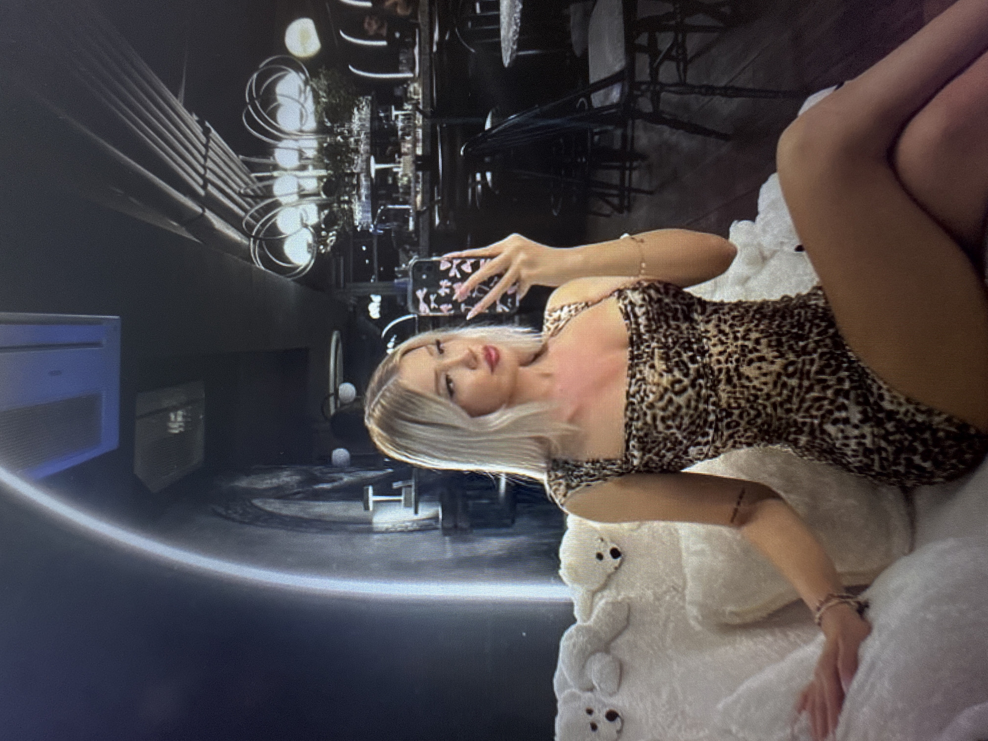 A young woman with straight, light-blonde hair and red lipstick is the central subject, reclining on a plush white surface, possibly a bed or large cushion. She is wearing a sleeveless, leopard-print mini-dress or romper. Her left hand is raised, holding a smartphone with a black case adorned with pink and white butterfly or floral patterns, suggesting she is taking a selfie or being photographed with the phone visible. Gold-toned bracelets are visible on both wrists.
The white surface she is on features several small, attached plush figures that resemble white bears with dark eyes and noses.
The background, visible as a reflection, appears to be a stylish indoor setting, likely a restaurant, bar, or lounge. It is dimly lit, featuring dark furniture such as tables and chairs. Distinctive lighting fixtures, characterized by glowing circular or spiral designs, are prominent throughout the reflected space. Potted greenery can also be discerned among the tables. On the far left of the background, a modern light-blue glowing panel or strip is visible, adding to the contemporary ambiance. The overall impression is one of an evening or nighttime setting. There is no discernible text in the image.