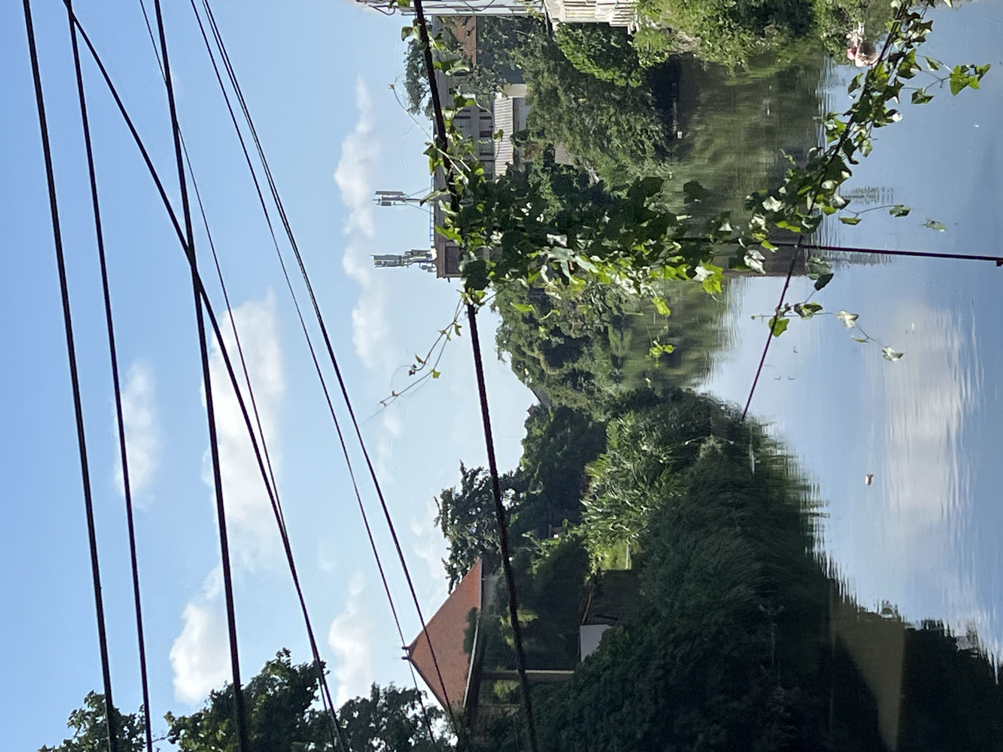 The image captures a tranquil view of a river flowing through a densely vegetated urban area, possibly in Kuta, Indonesia. Overhead, a complex web of dark electrical wires crisscrosses the sky, partially obscuring the bright blue sky dotted with scattered white clouds. The riverbanks are lined with lush green foliage, including trees and tall grasses, with some areas showing evidence of human structures. On the left bank, a building with a distinctive terracotta-tiled, pyramid-shaped roof is visible, partially covered by ivy. Further in the distance, more buildings emerge from the greenery, including structures with prominent telecommunication towers. The water of the river is calm, reflecting the sky and the surrounding vegetation, with a small, light-colored object floating on its surface towards the right side of the frame. A vine with green leaves hangs down from the overhead wires in the foreground, adding a natural element to the scene. No people are visible in the image.