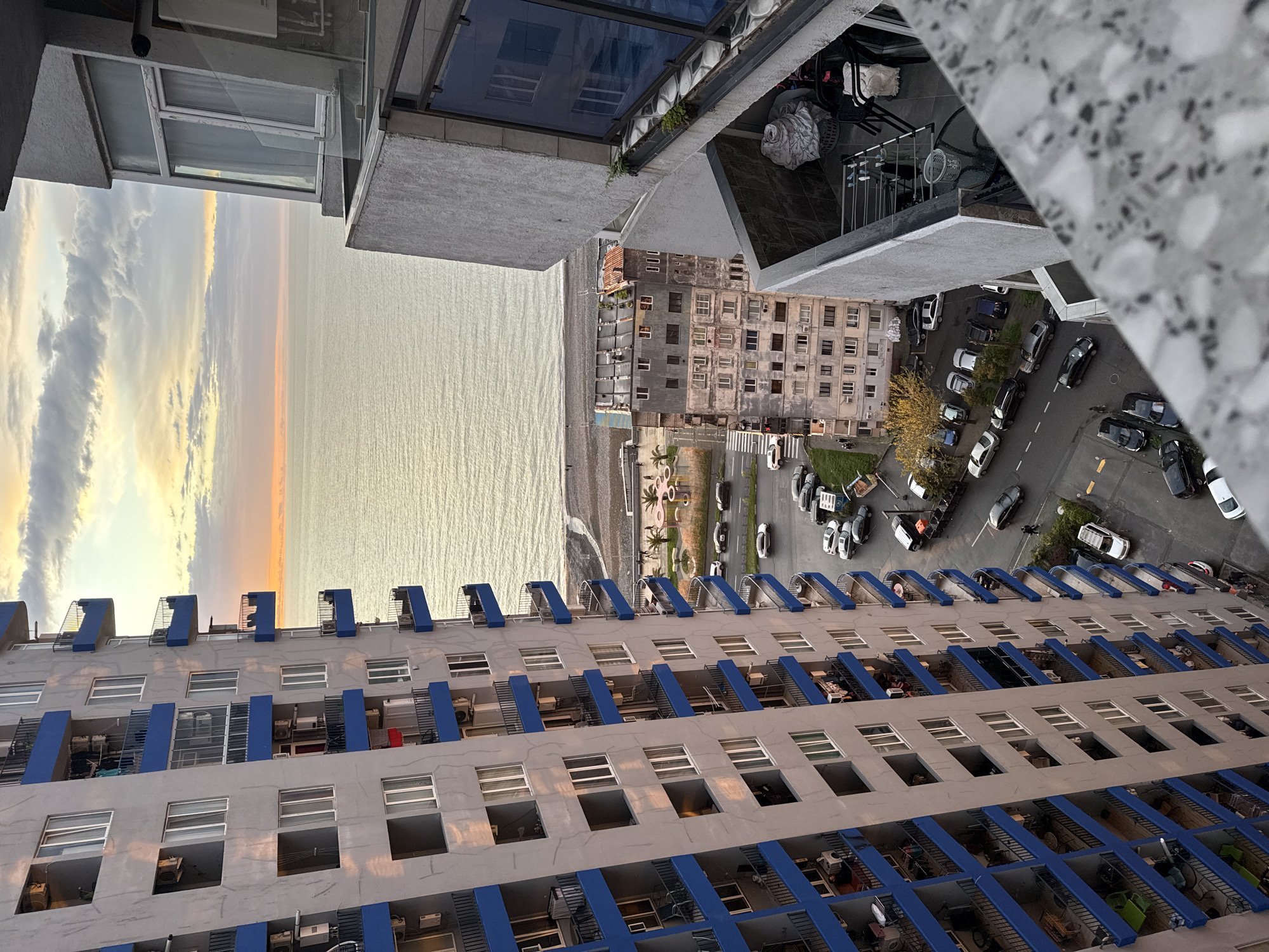 A high-angle perspective captures a cityscape bordering the sea at sunset. The image seems to be taken from a high-rise building, looking down at another building. This adjacent building, painted in gray with blue balconies, features rows of windows and balconies. Adjacent to the building is a beach area with parked cars, pedestrian areas, and another smaller residential structure. The sea stretches out to the horizon, reflecting the colors of the sunset sky above. The location is most likely Batumi, Georgia.