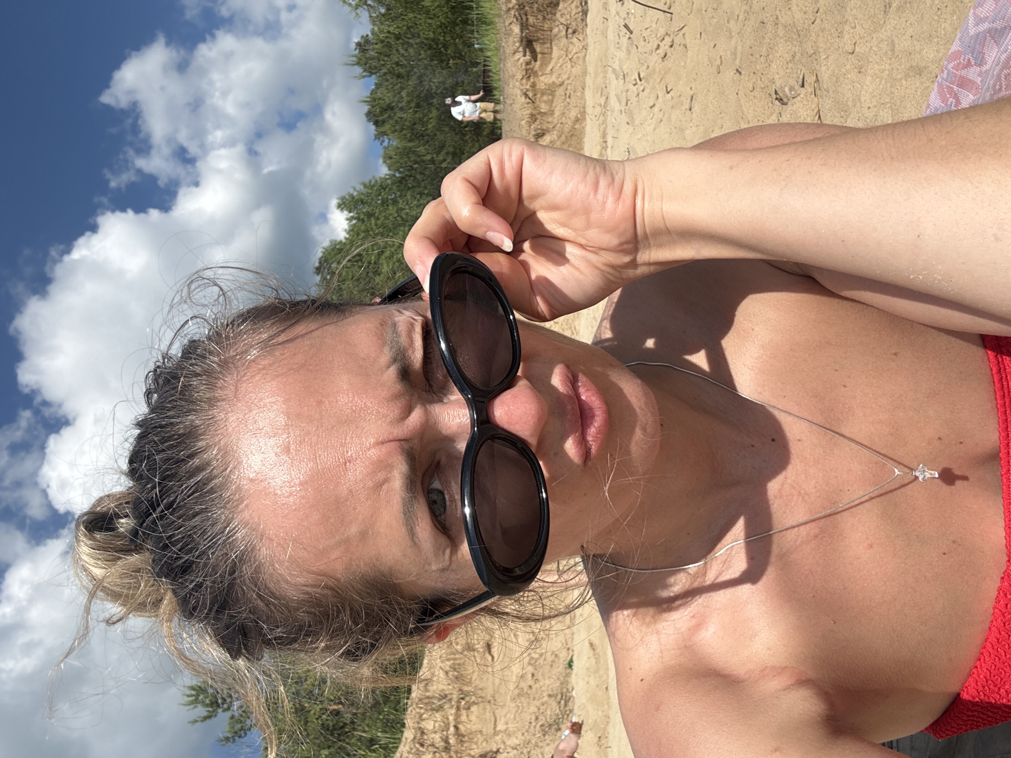 A medium shot captures a woman in the foreground, sitting on a sandy beach under a bright, cloudy blue sky. She is looking directly at the camera with a playful, squinting expression, her lips pursed. Her hair is pulled up into a messy bun, with loose strands framing her face. She wears black, oval-shaped sunglasses, which she is adjusting with her right hand, bringing them down slightly on her nose. A silver necklace with a small cross pendant is visible around her neck. She is wearing a red garment, likely a bikini top, and her bare shoulders and upper chest are exposed, showing some sun-kissed skin. The background depicts a sandy bank with dry grass and sparse trees. In the distance, another person, a man wearing a white t-shirt and shorts, is visible standing on the bank, seemingly engaged in an activity. The overall scene suggests a casual day at the beach. The location is identified as the city of Kzyl-Bairak, Russia.