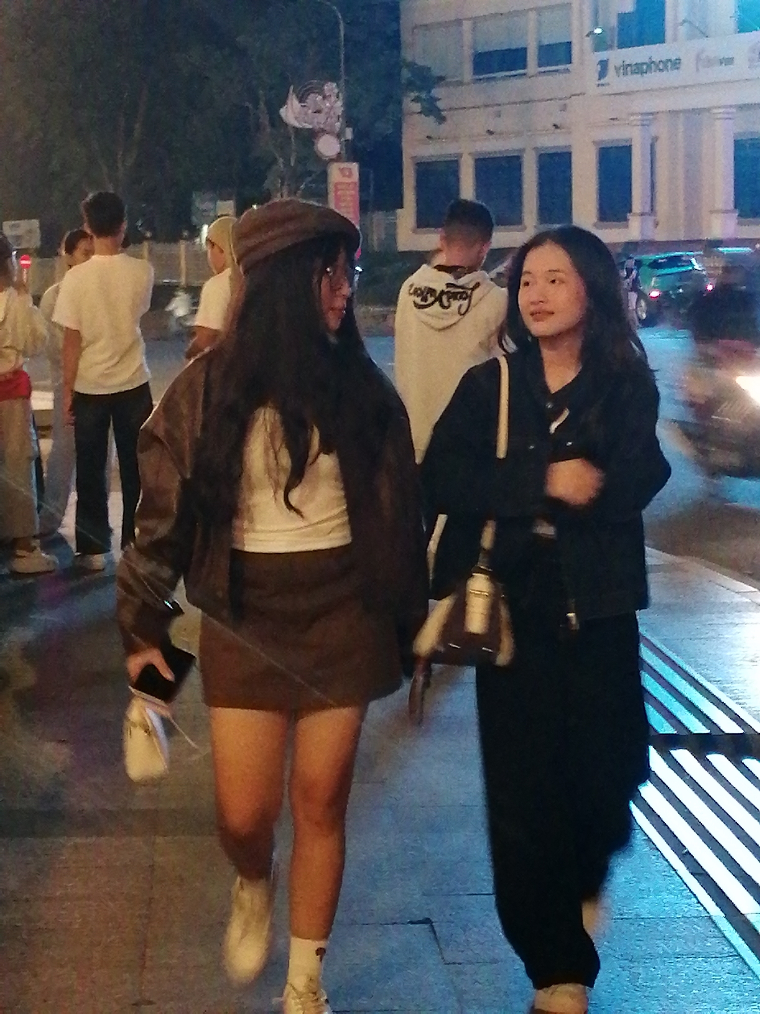 Two young women are walking on a sidewalk at night in Da Lat, Vietnam. The woman on the left is wearing a brown flat cap, glasses, a white top, a brown jacket, and a brown skirt. She is holding a black phone and a white purse. The woman on the right is wearing a black jacket and black pants. She is carrying a brown and white purse. In the background, other people are walking, and there are buildings and cars. The Vinaphone building is visible. The lighting suggests it is evening or night.