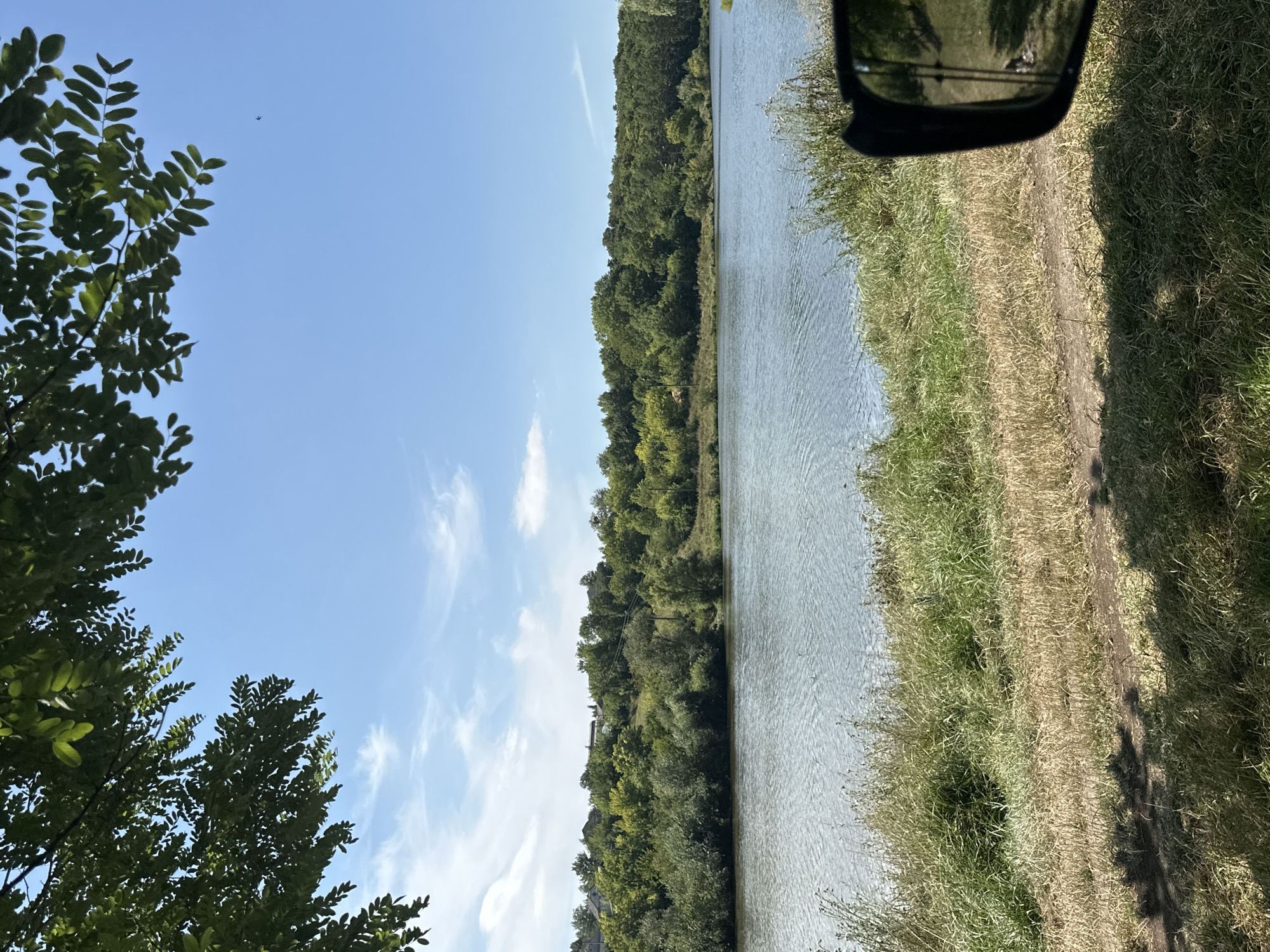 The image shows a scenic view of a calm lake with lush green trees and vegetation lining the opposite bank. The sky is a clear blue with a few wispy white clouds. In the foreground, dry and green grass covers the bank of the lake, with the shadow of tree branches cast upon it. A side-view mirror of a vehicle is visible on the right side of the frame, reflecting a patch of grass and sky. The scene appears to be located in the village of Makiv, Ukraine, capturing a peaceful natural landscape.