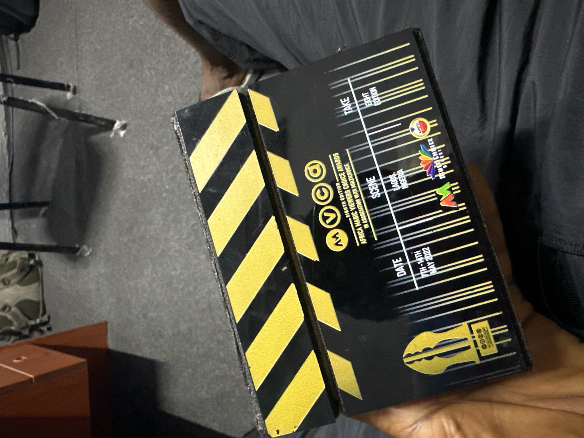 The image shows a black and gold clapperboard featuring details for the "Eighth Edition, Africa Magic Viewers Choice Awards" in association with Multichoice, taking place in Lagos, Nigeria, with the event dates of May 7th-14th, 2022. The clapperboard has a logo on it representing the awards as well as logos for Amstel and Multichoice. The item is being held by a person wearing dark clothing. The surrounding environment is blurry but includes a chair and a small table. Ikeja, Nigeria, is not mentioned on the clapperboard.