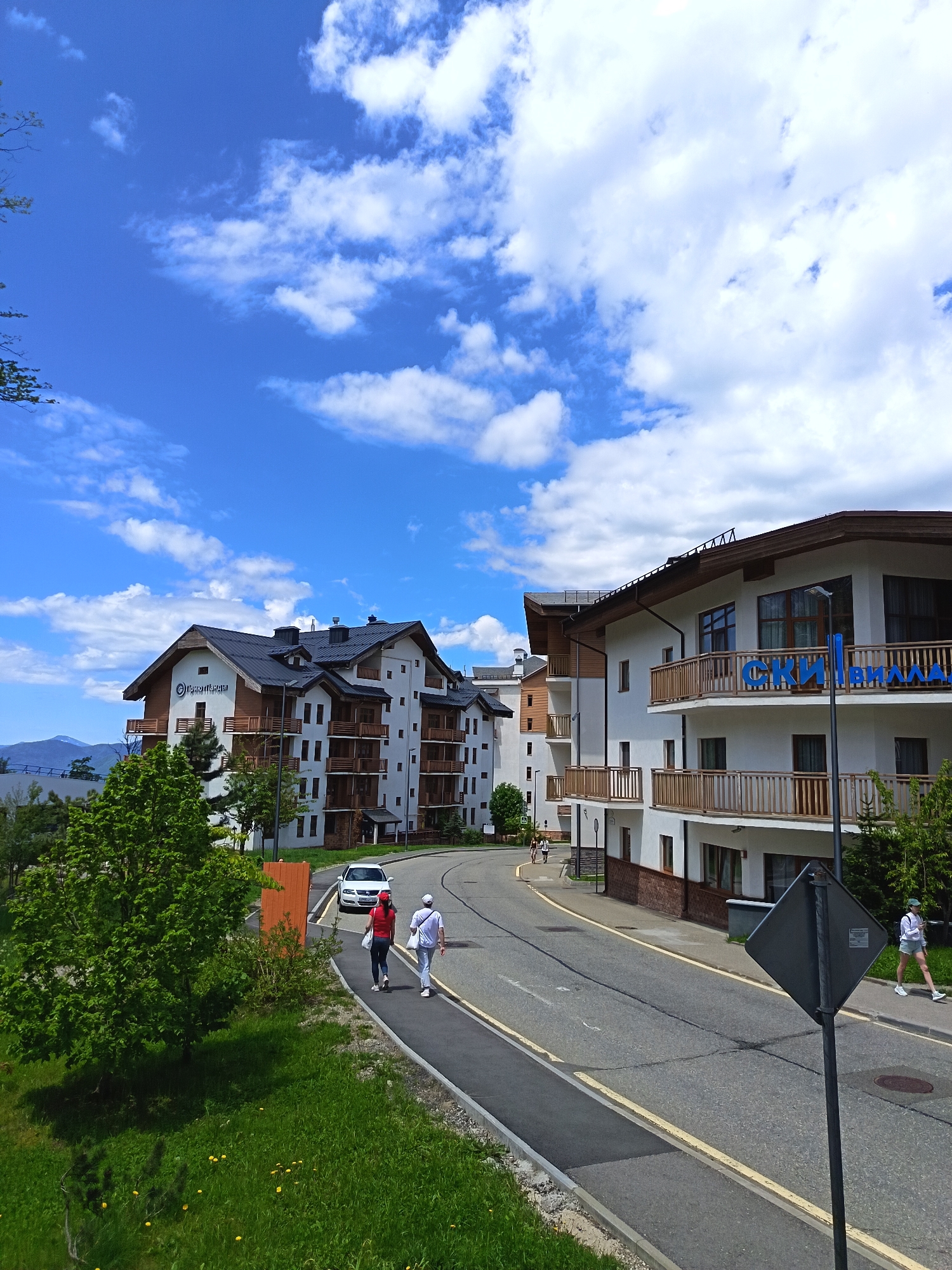 The image displays a sunny daytime view of a road winding through a resort area in Estosadok, Russia. The sky is a vibrant blue dotted with numerous white, cumulus clouds.
On either side of the asphalt road, modern multi-story buildings with white exteriors, dark roofs, and wooden balconies stand. The building on the left features a sign with Cyrillic text that reads "ПолянаСказы" (PolyanaSkazy). On the right, another building has a large blue sign spelling "СКИ|ВИЛЛА" (SKI|VILLA). The architecture suggests an alpine or mountain resort style.
Several people are visible walking along the sidewalks. In the mid-ground, two individuals, one in a red top and another in white, walk away from the camera. A white car is parked near the sidewalk next to them, partially obscuring its license plate, which shows "P833AP 93". Further down on the right, another person is walking towards the camera.
The road has yellow lane markings and exhibits some wear with minor cracks. Lush green grass and various trees line the sides of the road, and dandelions are visible in the foreground grass. In the far distance, faint mountain ranges can be seen beneath the clear horizon. A dark, square road sign, viewed from the back, is prominent in the lower right foreground.