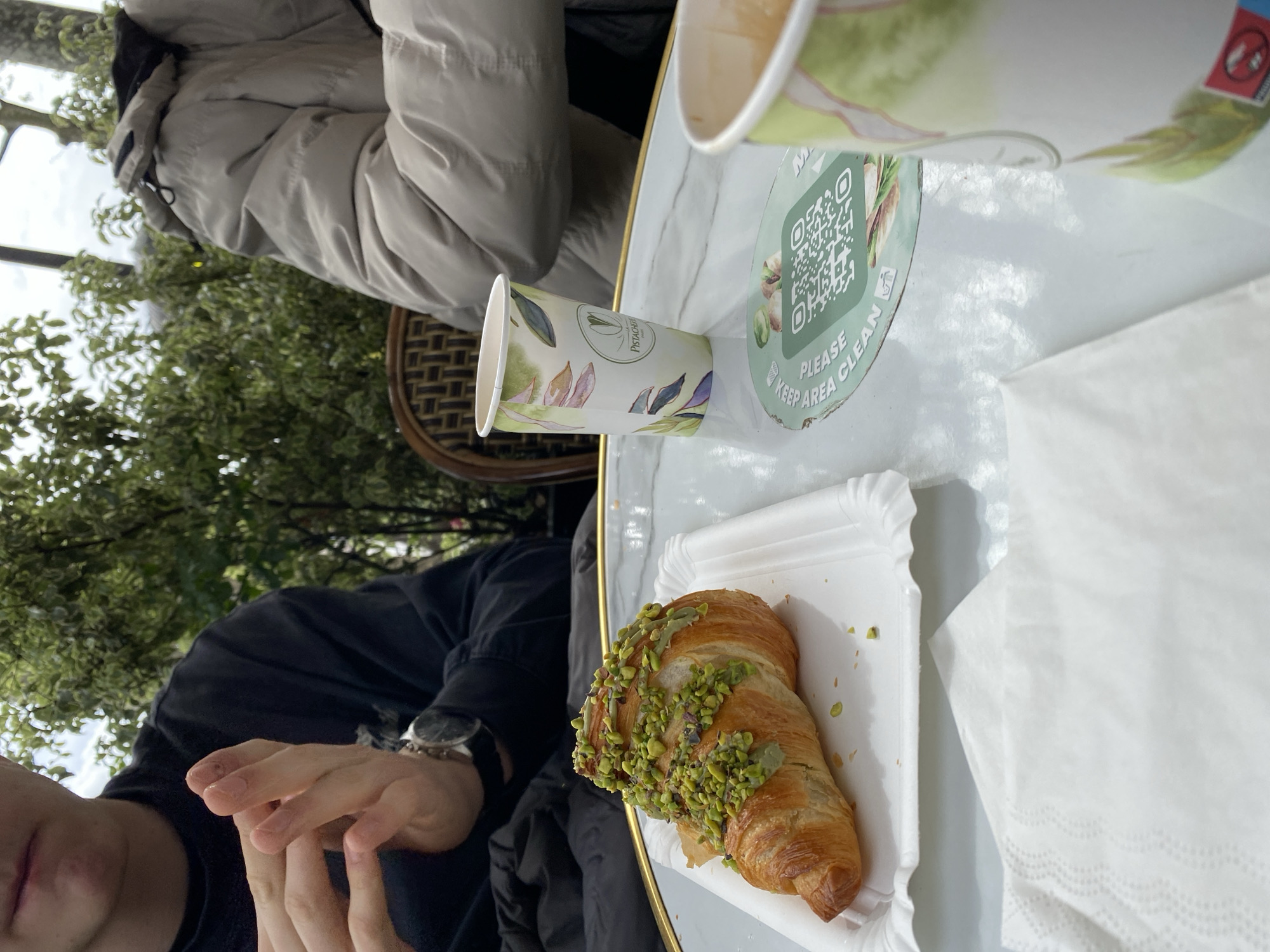 The image shows a scene at a café, likely in Paris, France, based on the presence of a "Pistachio" brand cup, a pastry dusted with green nuts (likely pistachios), and French café-style seating in the background. Two individuals are partially visible: one wearing a beige puffer jacket and another in a black top. A paper cup and a larger cup, possibly containing a beverage, sit on the white table. The pastry, presumably a croissant, sits on a small white paper plate next to a stack of napkins. The table has a sticker displaying a QR code. Outdoor trees are visible in the background.