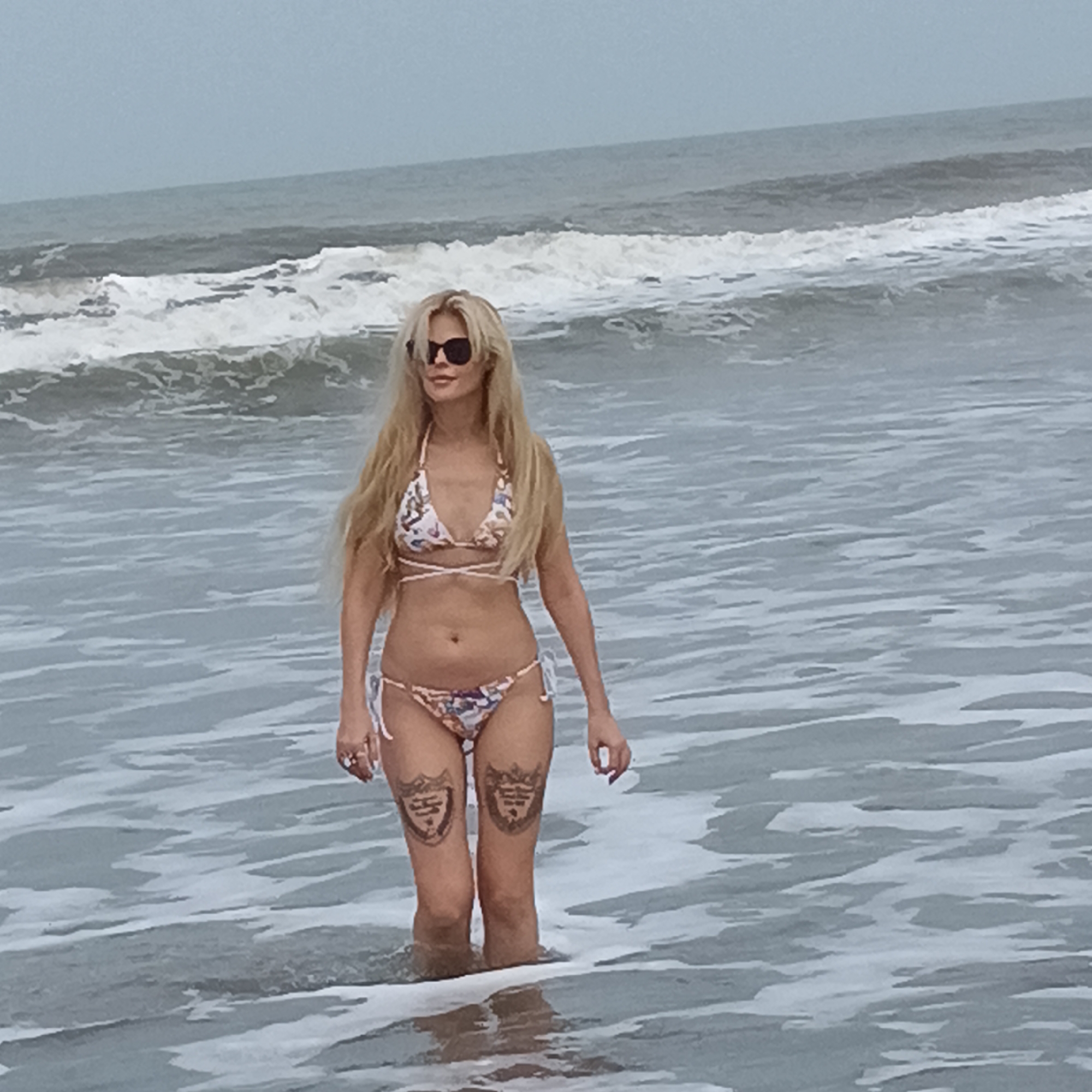 A blonde woman wearing a bikini and sunglasses stands in ocean water up to her lower legs. The woman has tattoos on the front of both thighs. The ocean has visible waves. The sky is overcast. Location is unknown.