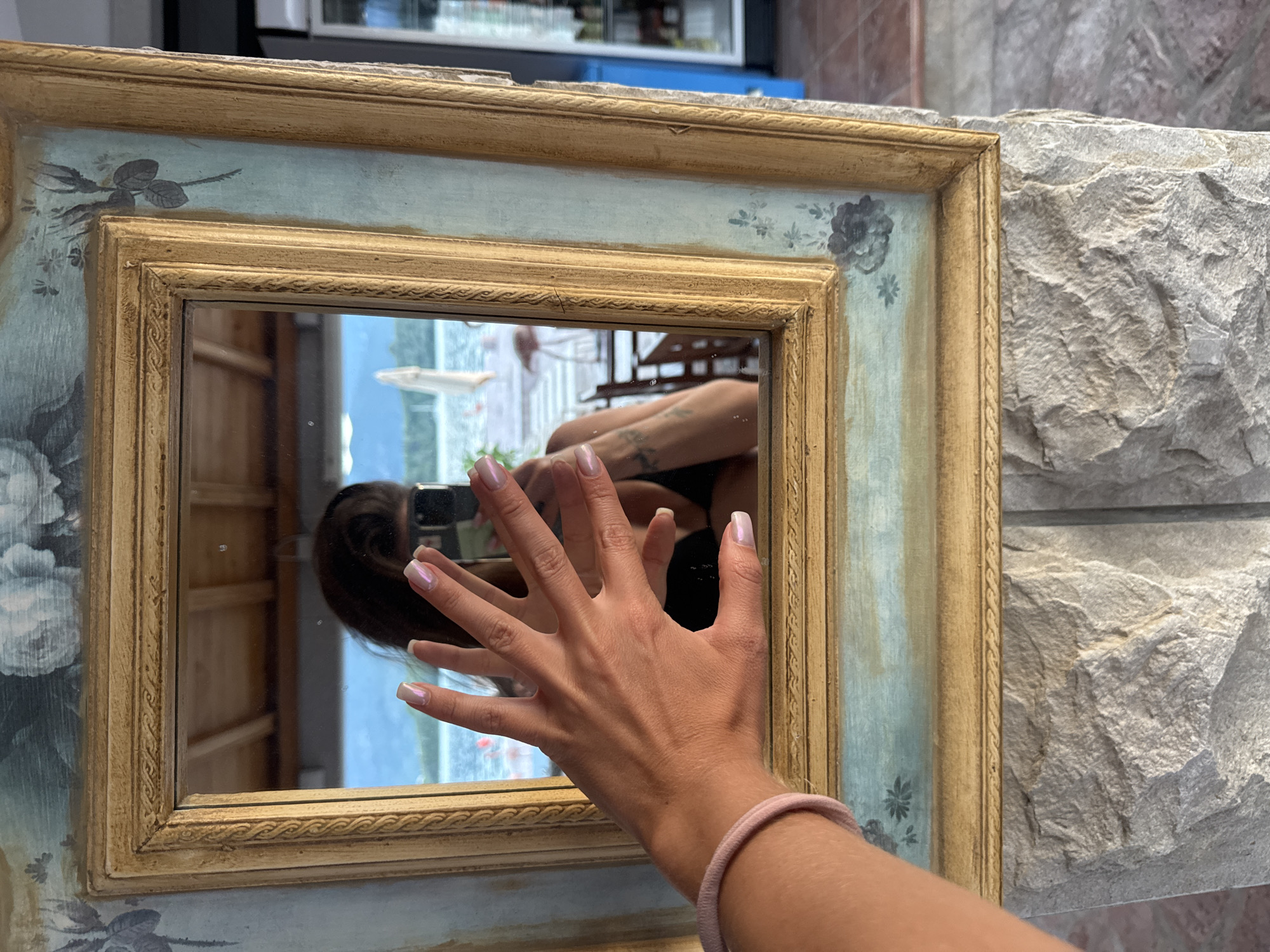 The scene depicts a framed mirror resting against a stone wall. The mirror's frame is ornate, featuring a light blue and gold color scheme with floral designs. Reflected in the mirror is a person holding a phone, likely taking a photo. Their hand with manicured nails is prominently displayed in the foreground. The background reflection shows an outdoor scene with glimpses of greenery and a body of water. Based on the prompt, this scene is located in Bogišići, Montenegro.