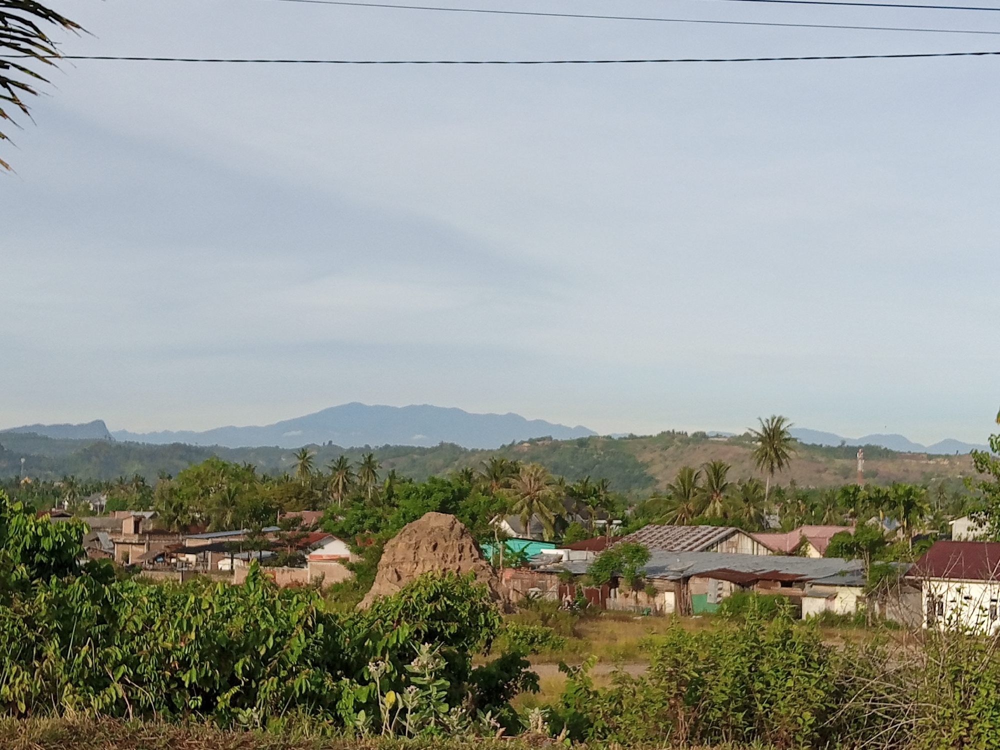 The image depicts a rural village scene in Uruekanoe, Indonesia. The foreground is filled with dense green foliage and shrubs, with a dry, rough patch of ground in the very bottom. Behind the foliage, a collection of modest houses with corrugated tin roofs and some with concrete walls are visible. The architecture suggests a tropical or sub-tropical environment. A large, rough mound of earth or rock formation stands out in the mid-ground, near some of the houses. In the background, lush green hills are covered with trees, and further beyond, a range of distant mountains under a pale, hazy sky. There are several utility wires stretched across the top of the frame. No people are present in the image.