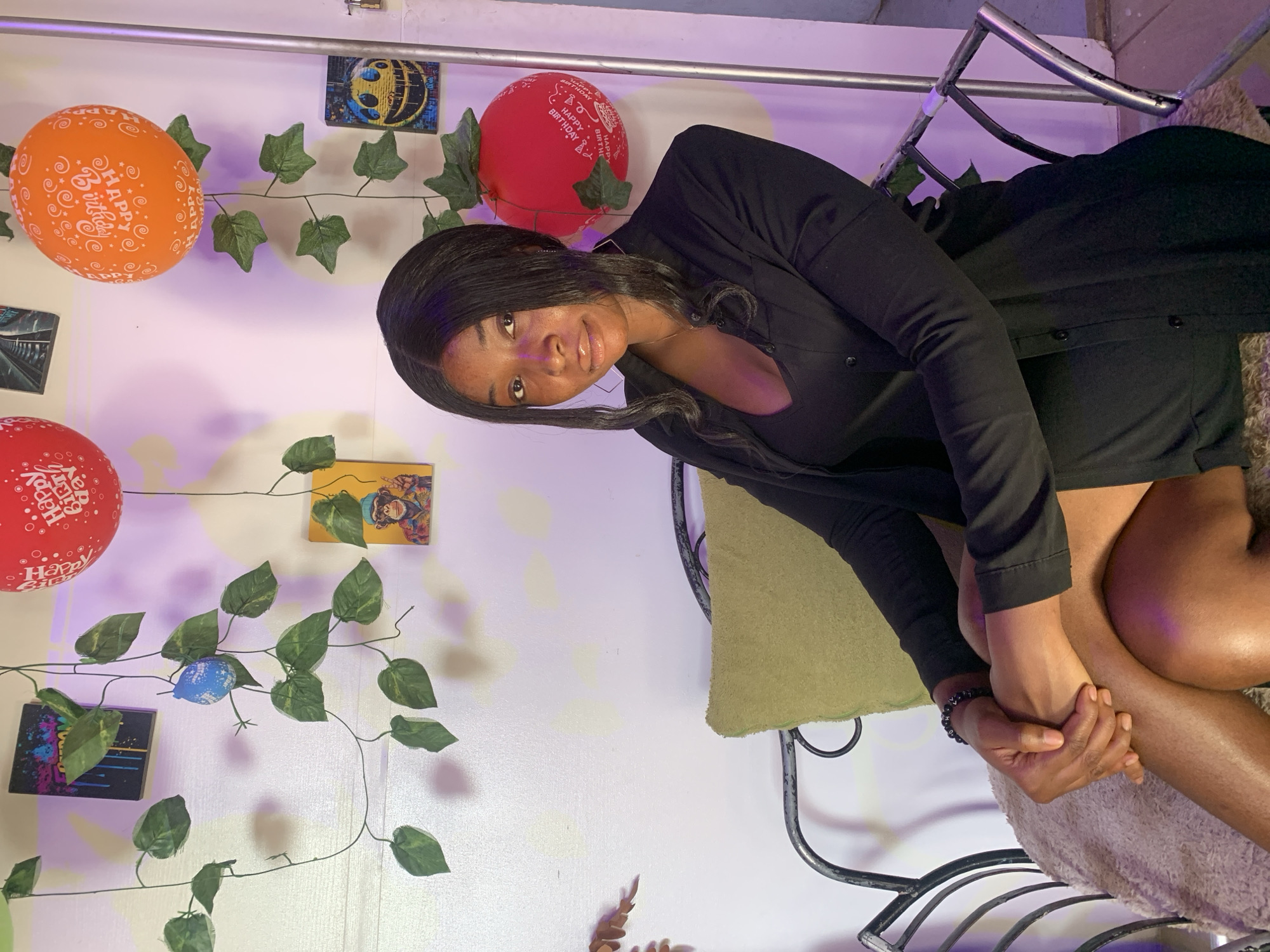 A young woman with dark hair and a black outfit sits on a chair with a green cushion. She has her hands clasped together. The background includes a white wall decorated with balloons that read "Happy Birthday", artificial ivy, and various small pictures. The setting appears to be indoors. The image does not contain specific indicators that place the location in Jos, Nigeria.