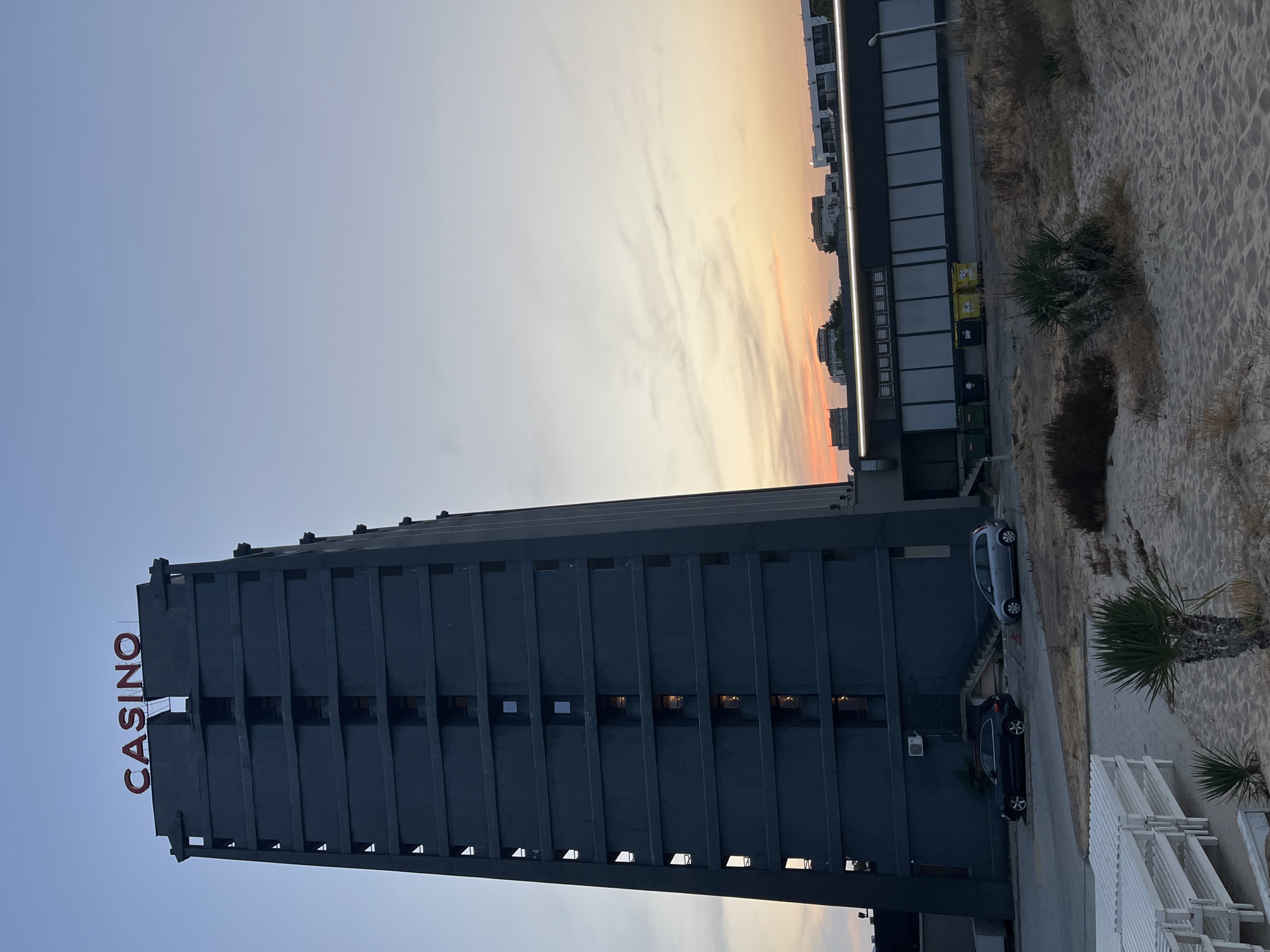 The image depicts the Casino building in the city of Nesebar, Bulgaria, at dusk. The tall, dark grey building dominates the frame, with the word "CASINO" in red lettering visible on its rooftop. The building features numerous dark, rectangular windows arranged in vertical rows. The sky above is a gradient of pale blue to soft orange and pink, indicating the setting sun, with faint clouds present.
In front of the Casino building, a long, dark-colored structure with large, reflective windows runs horizontally. Several colorful trash bins (green and yellow) are lined up against this structure. Two cars are parked on the paved area in front of the building: a dark SUV on the left and a silver minivan on the right.
The foreground of the image shows a sandy beach with several white, slatted lounge chairs stacked in the lower left corner. Sparse, dry vegetation and a small palm tree are scattered across the sand. The overall impression is a coastal urban scene during the transition from day to night.