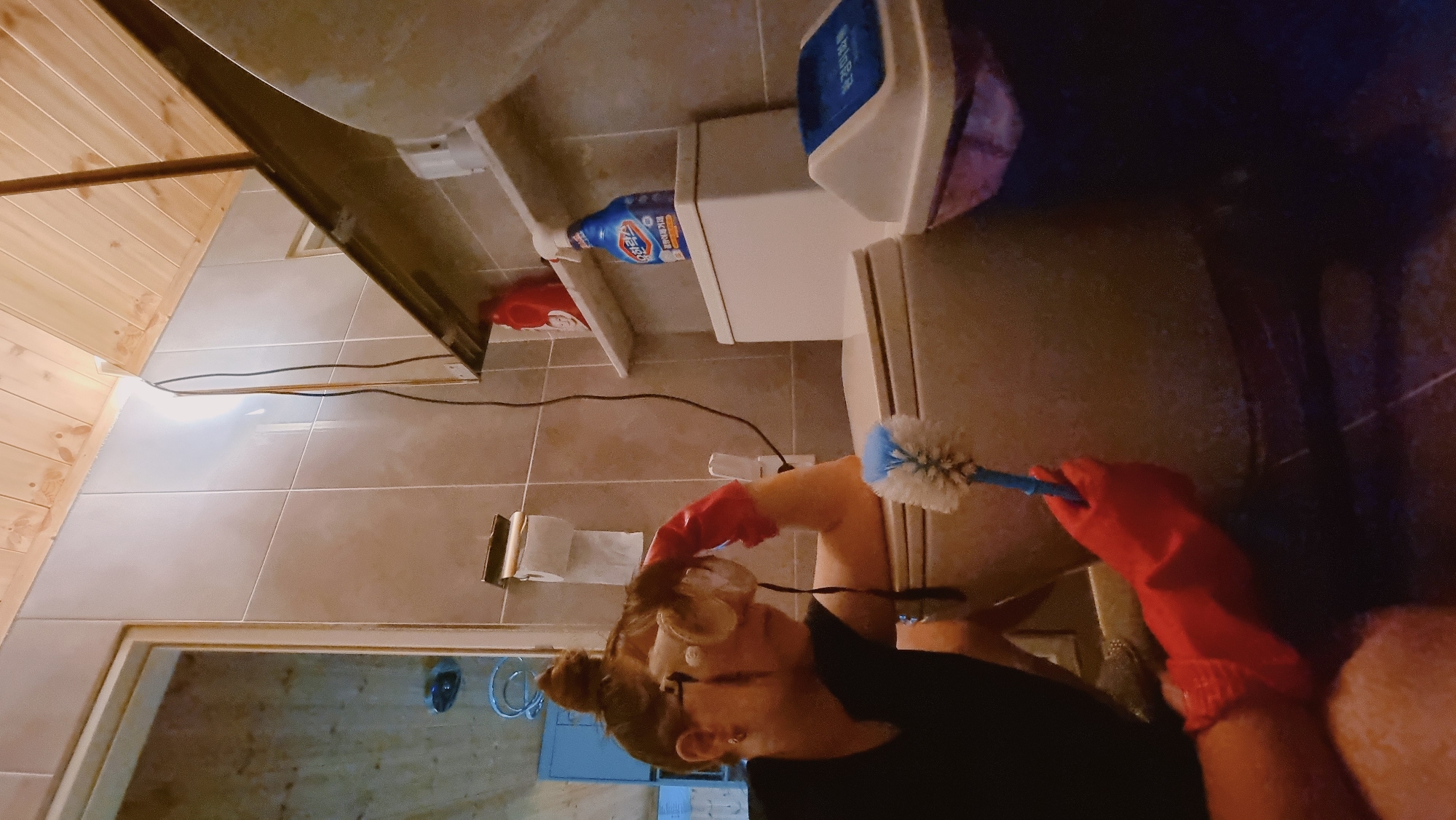 A woman in red gloves and safety glasses is cleaning a toilet with a blue and white toilet brush. The toilet is white and has cleaning supplies on a shelf above it. The bathroom has light brown tiles and a wooden ceiling. The location is Dunnae-myeon, South Korea.